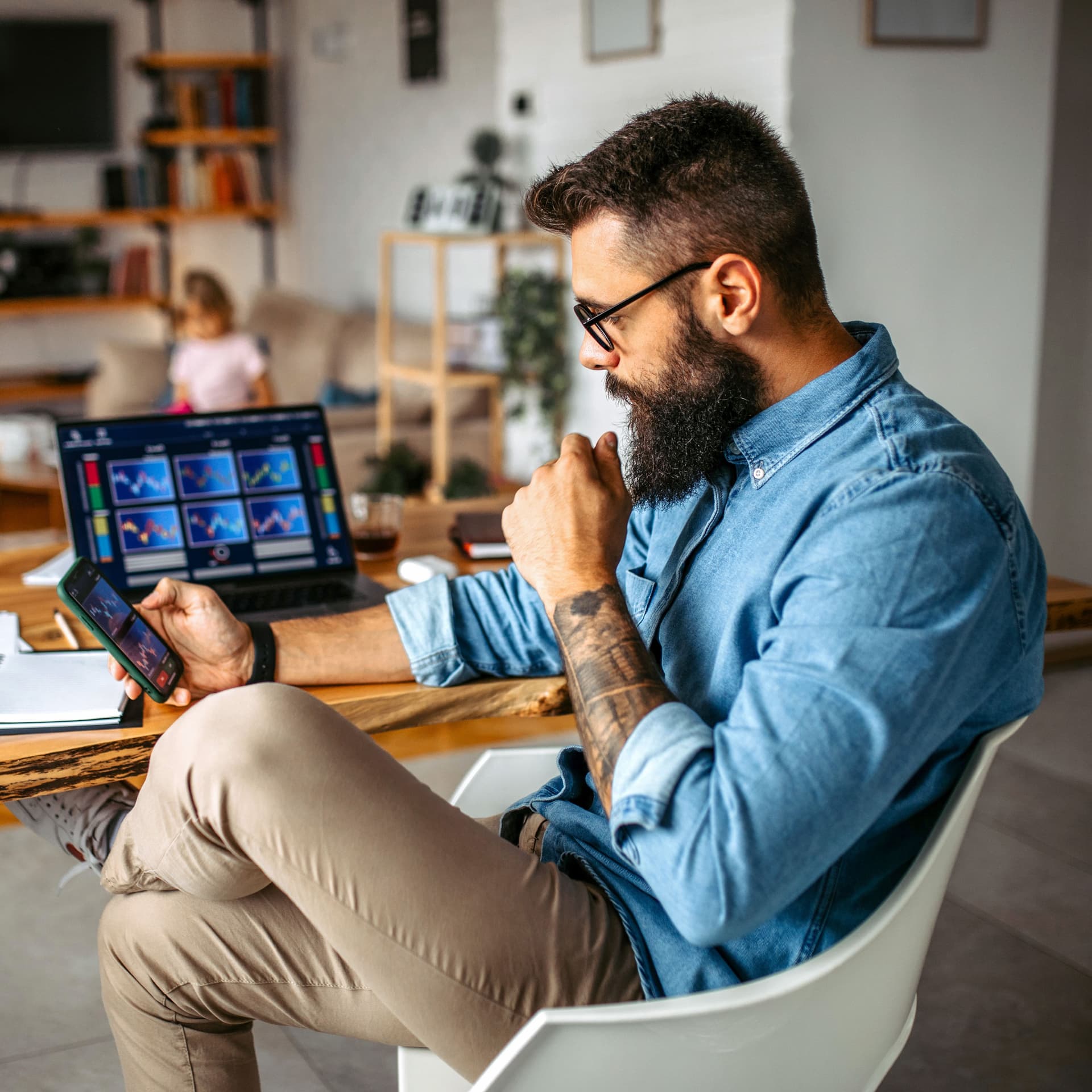 Mann sitzt am Schreibtisch im Homeoffice und blickt auf sein Smartphone, ein Laptop mit Diagrammen steht vor ihm.