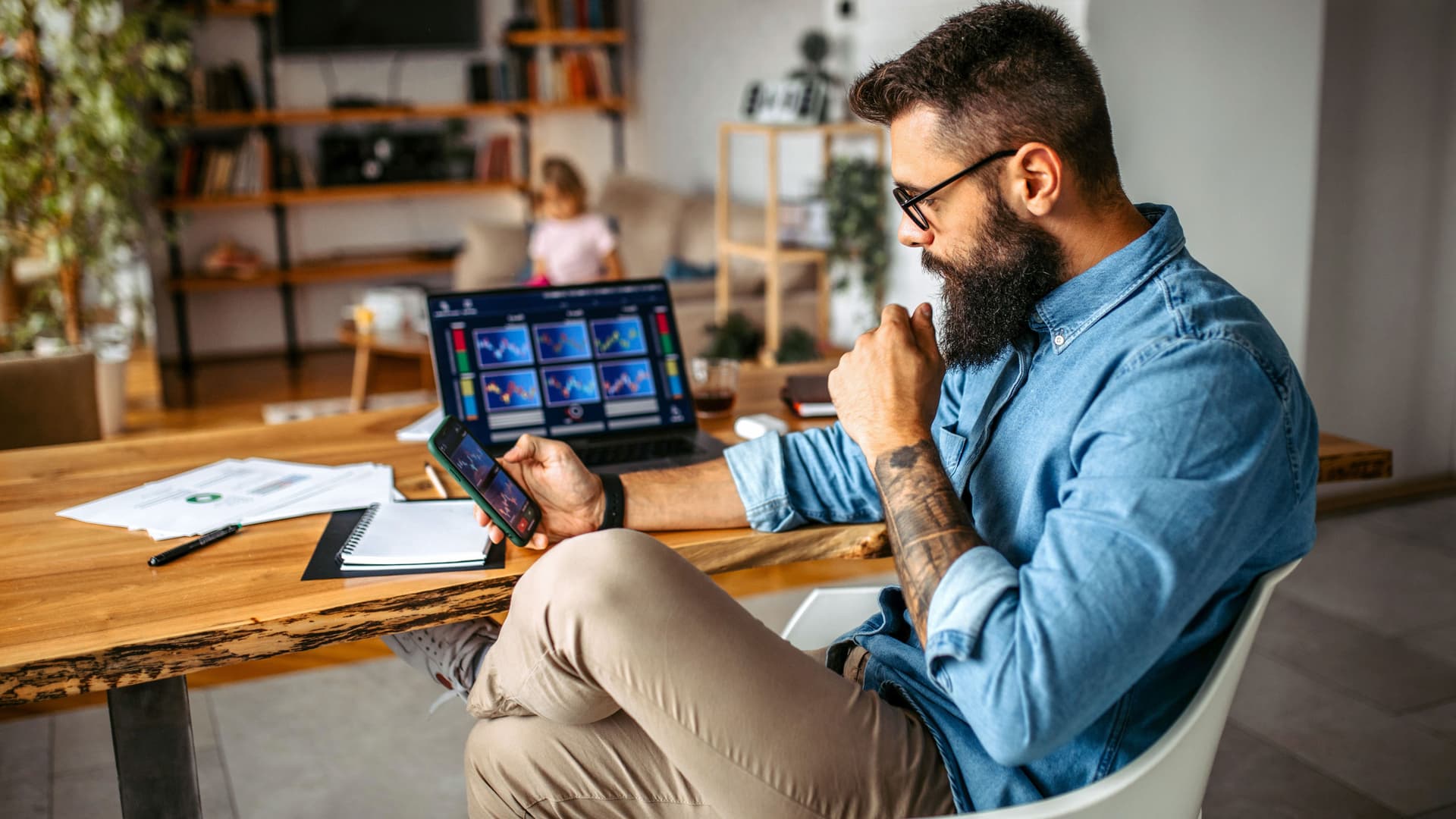 Mann sitzt am Schreibtisch im Homeoffice und blickt auf sein Smartphone, ein Laptop mit Diagrammen steht vor ihm.