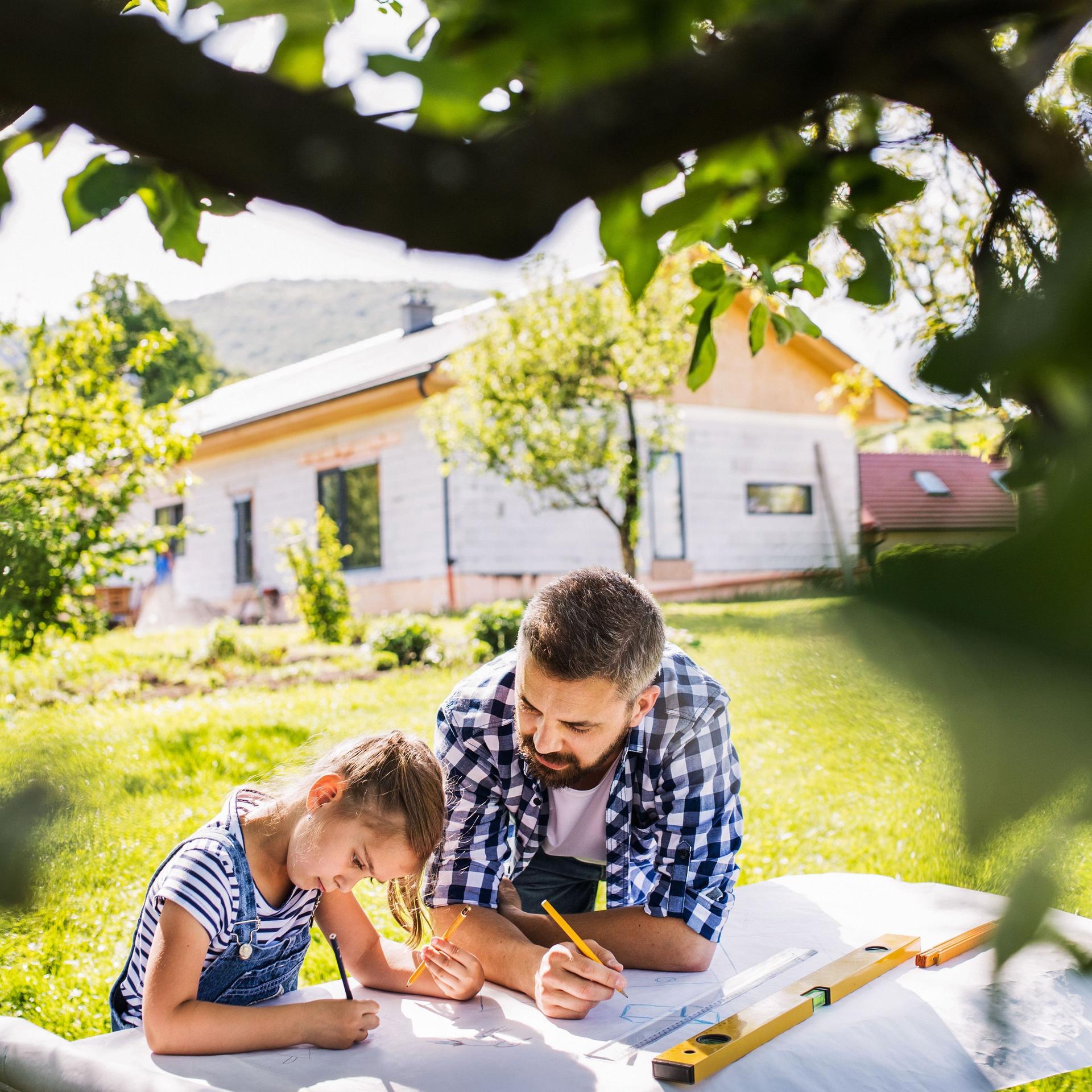 Vater und Tochter planen etwas im Garten mit Stift und Papier. Im Hintergrund steht ein Haus und im Vordergrund ein Baum.