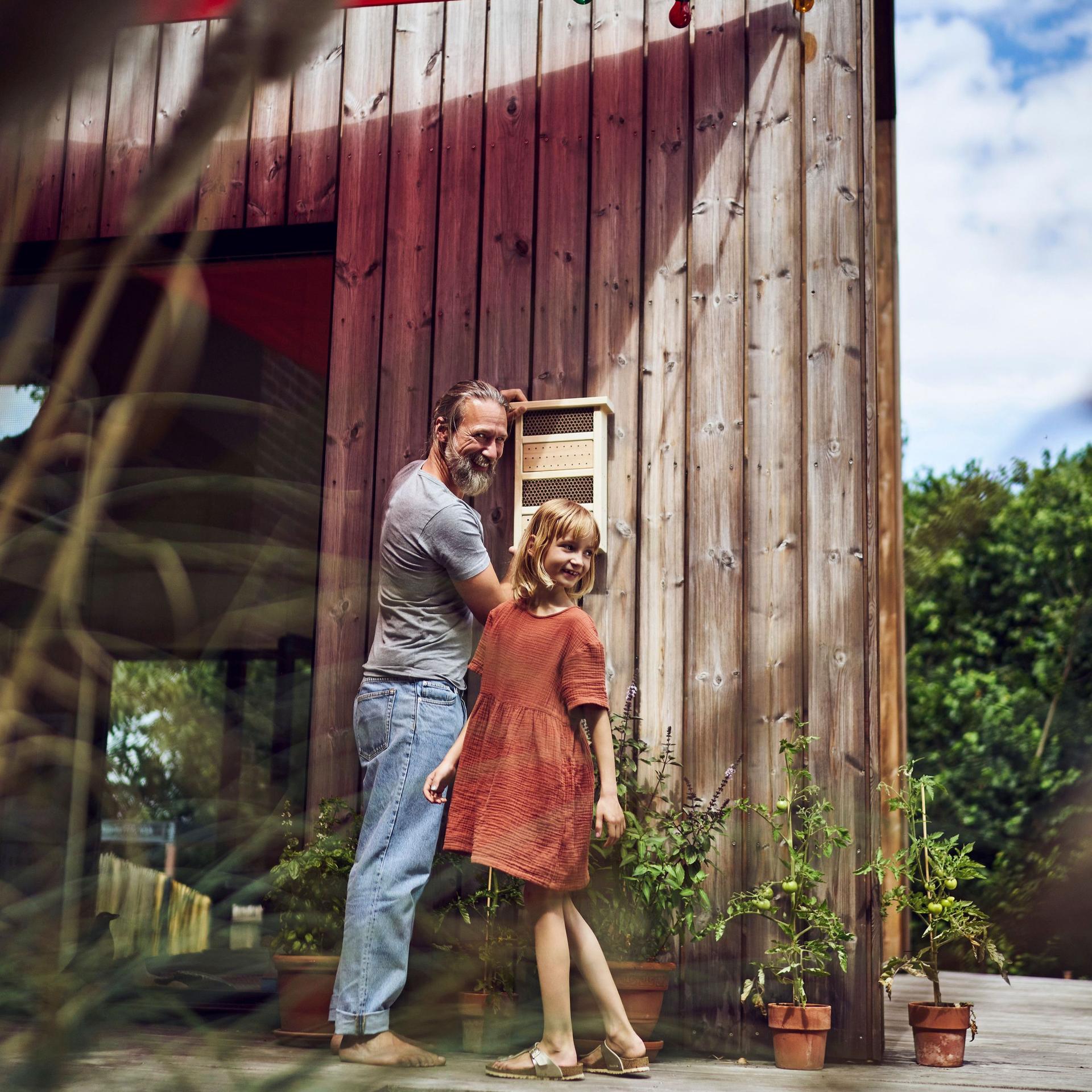 Vater und Tochter hängen ein Insektenhotel auf an ihrem Tinyhouse im Grünen.