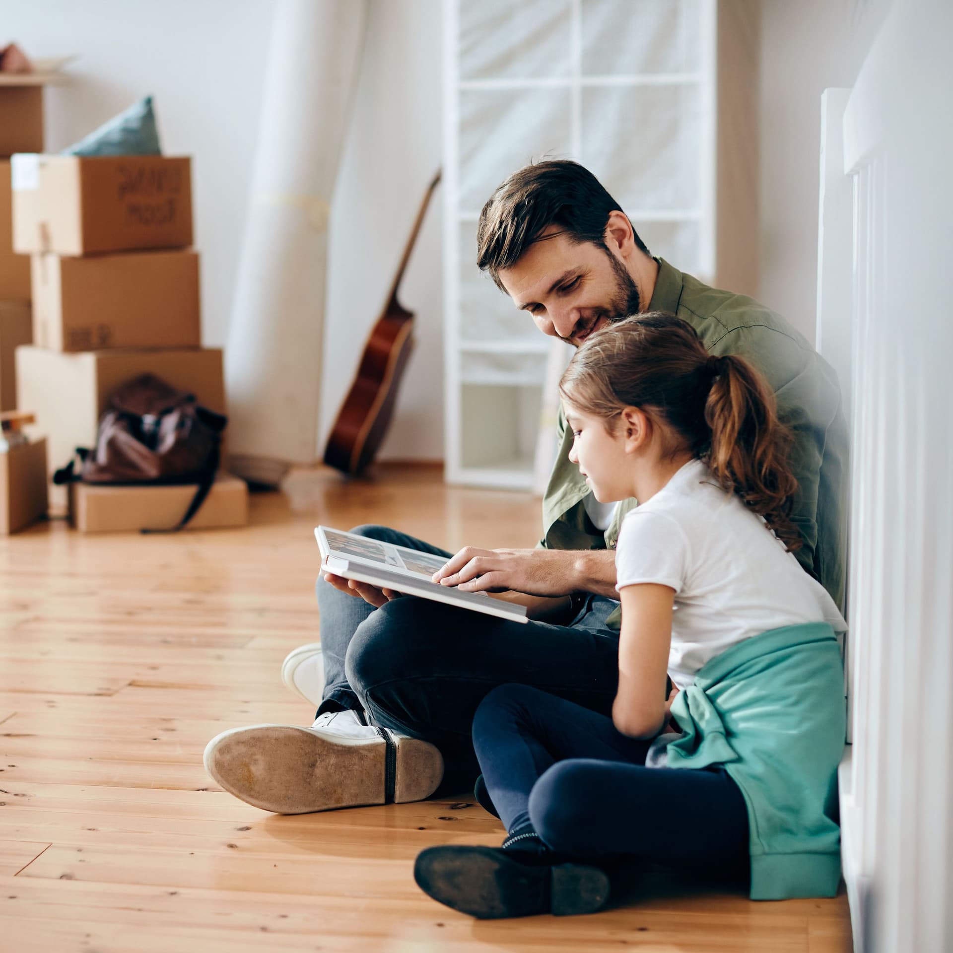 Lächelnder Vater und kleine Tochter sitzen auf dem Holzboden und gucken ein Fotoalbum an. Der Raum ist fast leer und im Hintergrund sind Umzugskisten.
