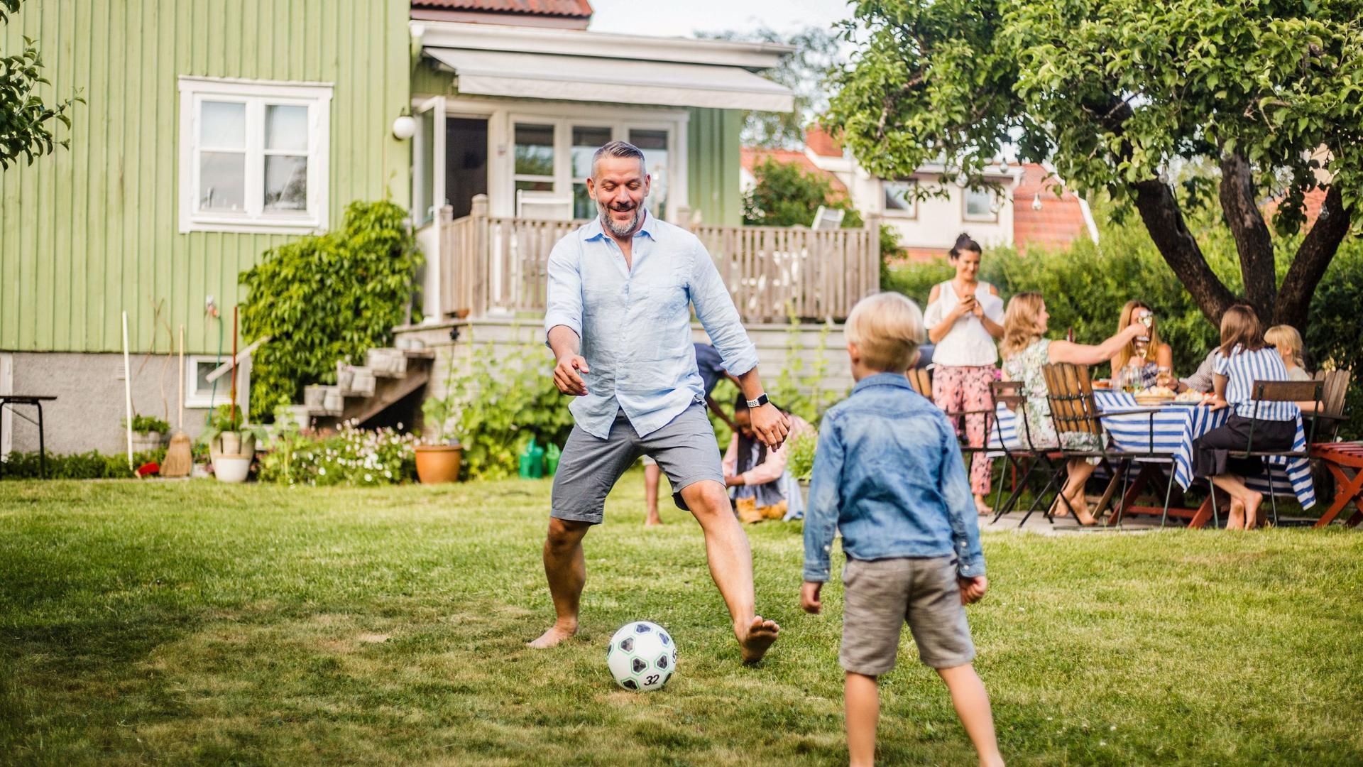 Vater spielt mit seinem Sohn im Garten eines Einfamilienhauses Fussball während im Hintergrund am Tisch gegessen wird.