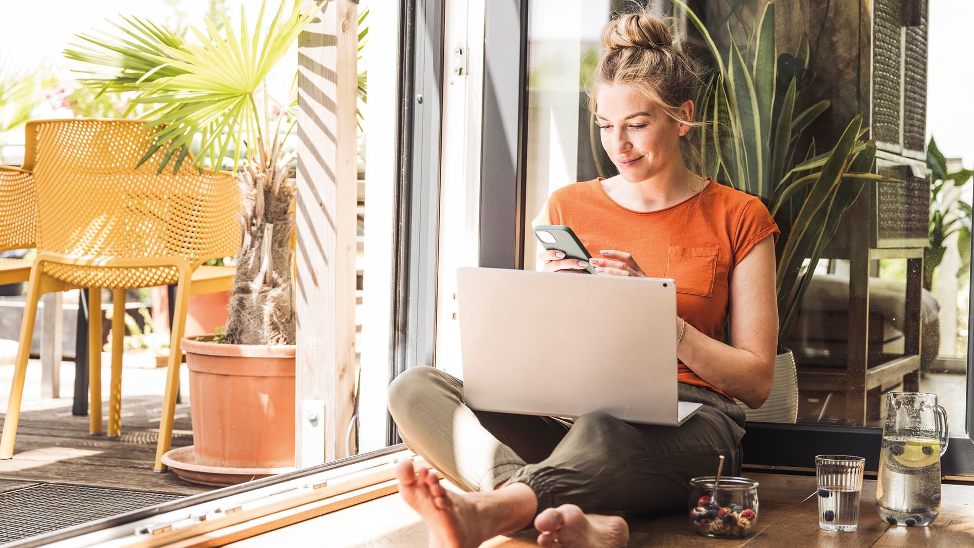 Eine junge Frau sitzt barfuß im Schneidersitz an einer geöffneten Terrassentür, hat einen Laptop auf dem Schoß und schaut lächelnd auf ihr Smartphone.