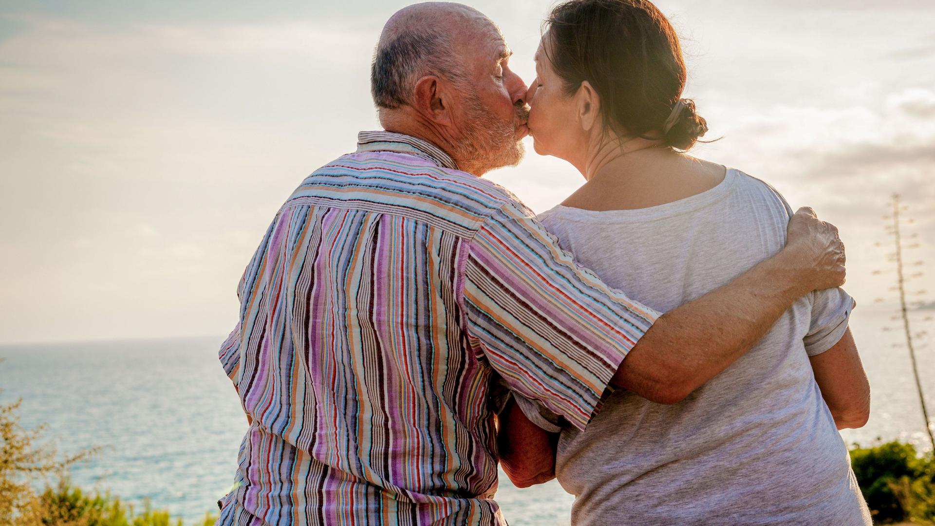 Zwei Senioren sitzen am Strand. Der Mann küsst seine Partnerin und umarmt sie dabei.