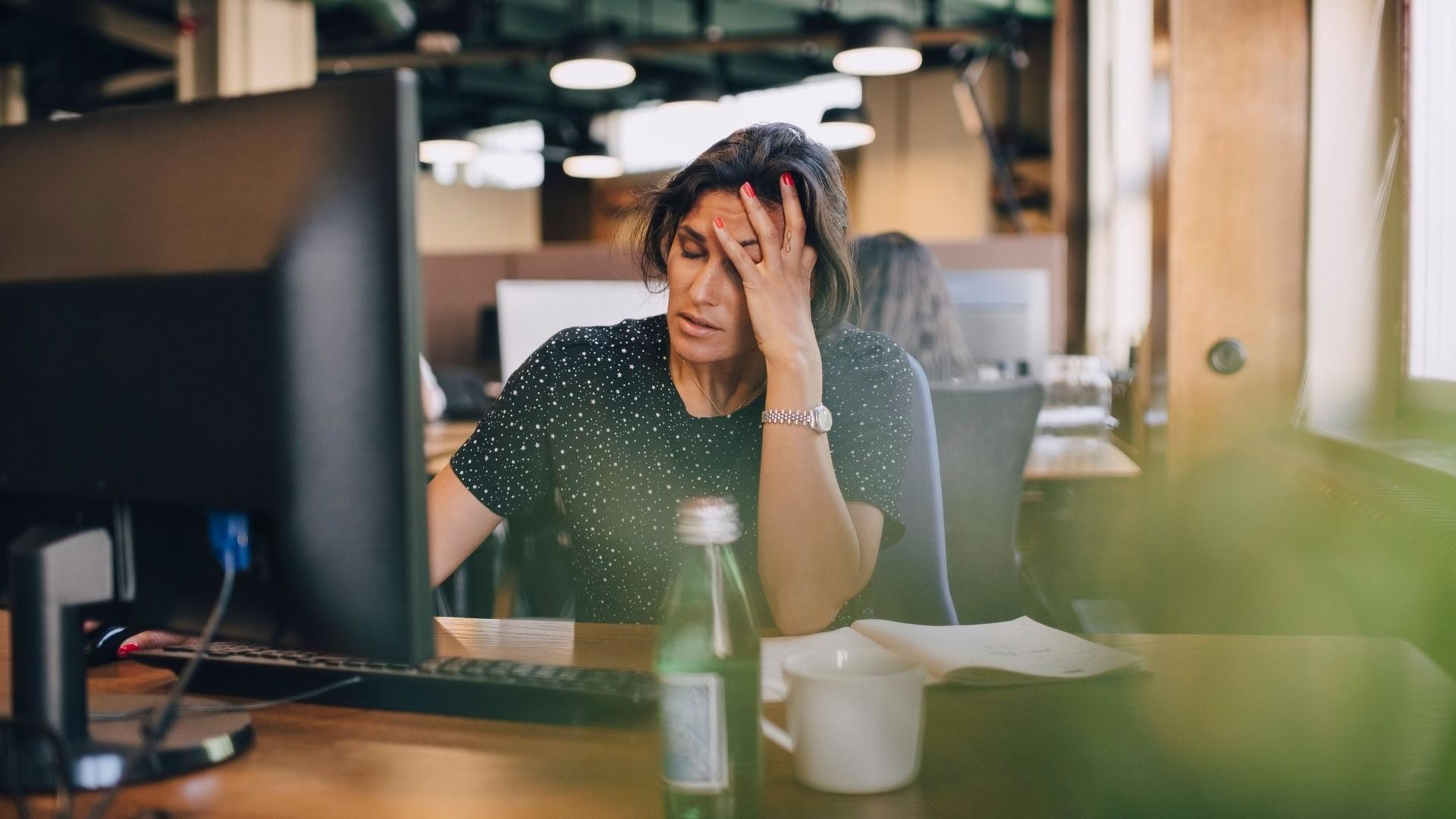 Eine Frau sitzt an einem Schreibtisch in einem Büro vor einem Computer und hält ihre Hand an den Kopf. Sie hat die Augen geschlossen und wirkt erschöpft.