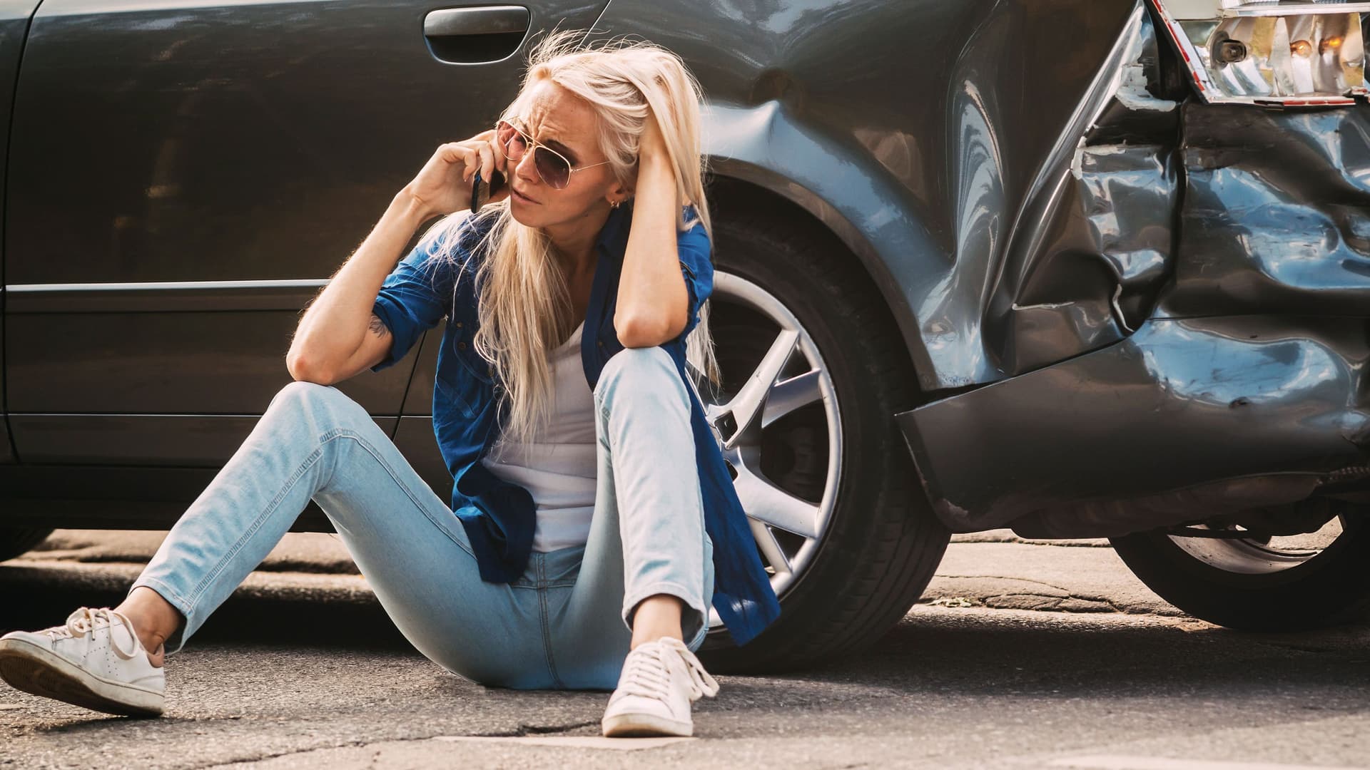 Frau sitzt an Unfallauto gelehnt und telefoniert. Das Heck des Autos ist verbeult.