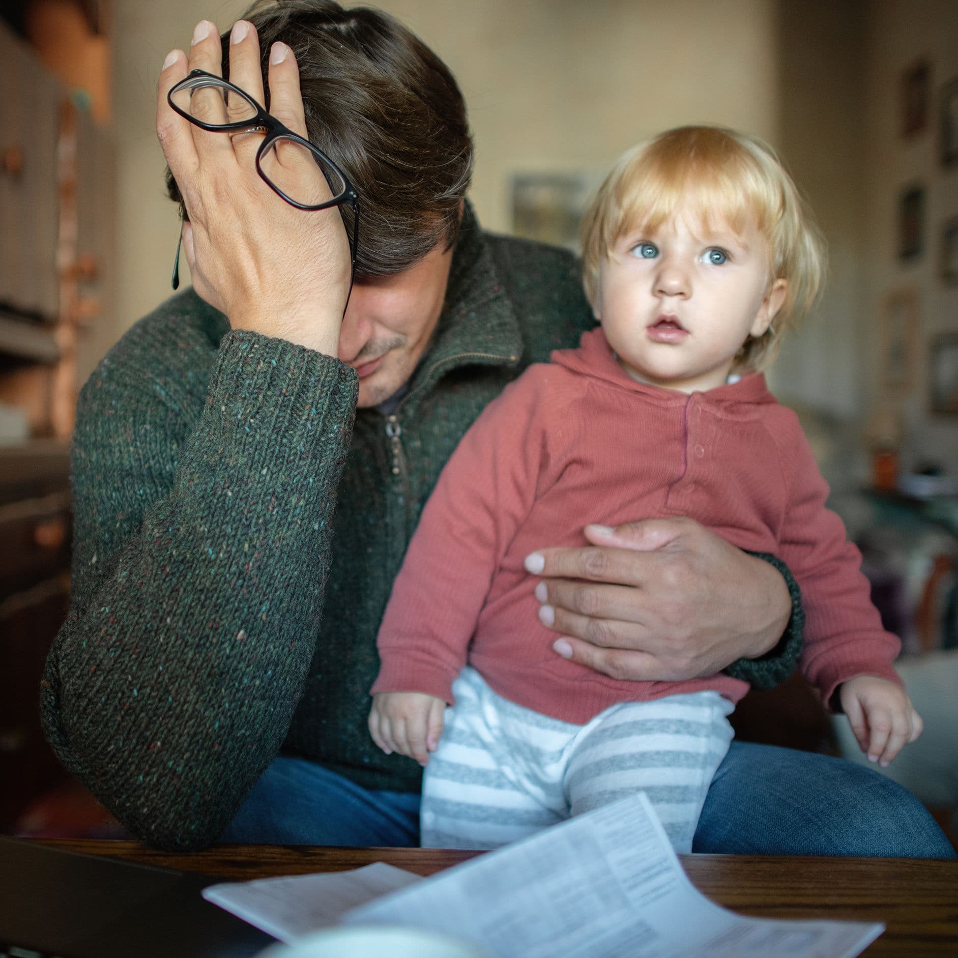 Ein Mann hält sich verzweifelt das Gesicht. Auf seinem Schoss sitzt ein Kleinkind. Auf dem Tisch liegen Rechnungen.