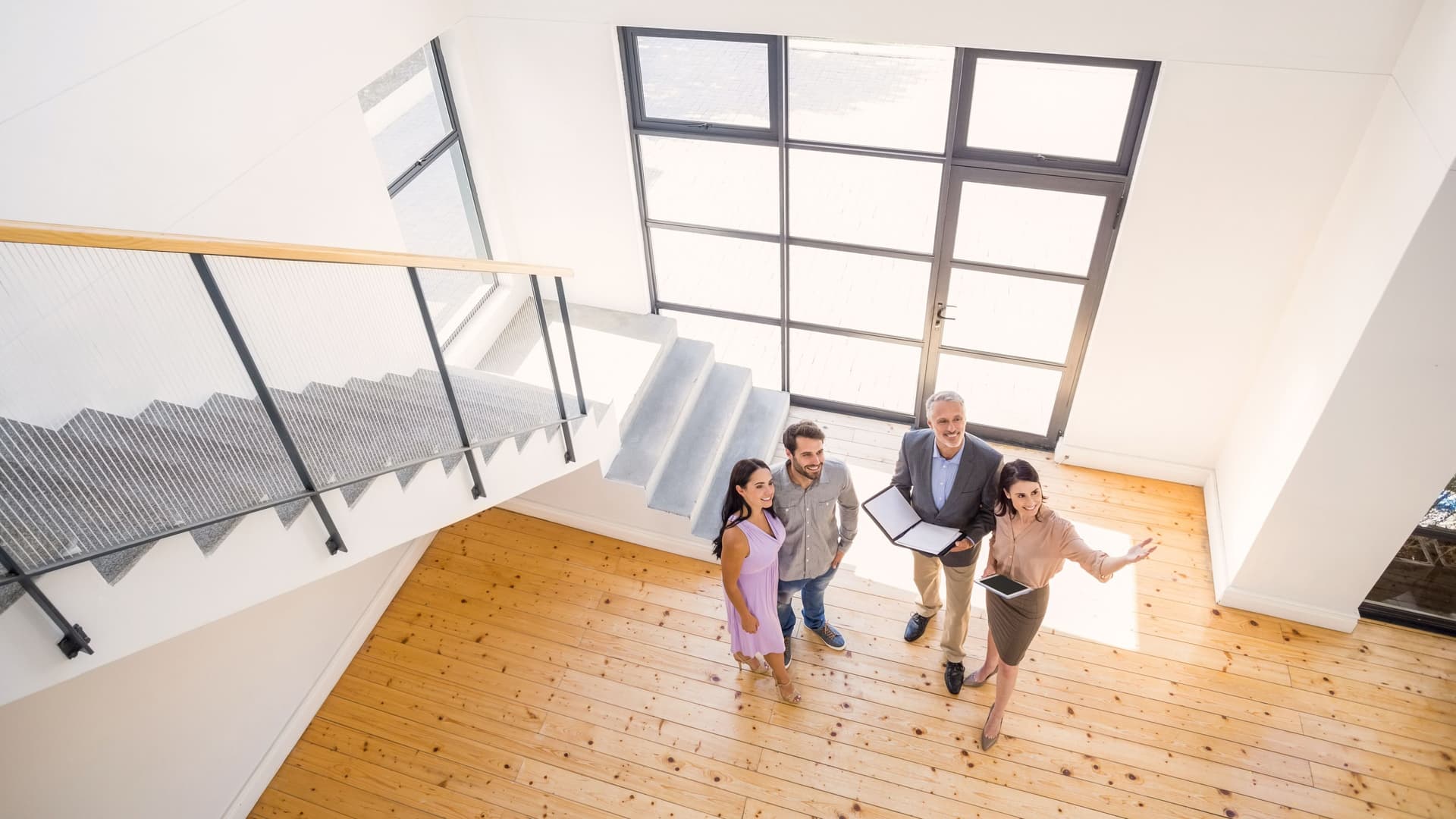 Vogelperspektive einer Maklerin mit drei Immobilienintressenten. Sie steht in offener und moderner Wohnung und gestikuliert.