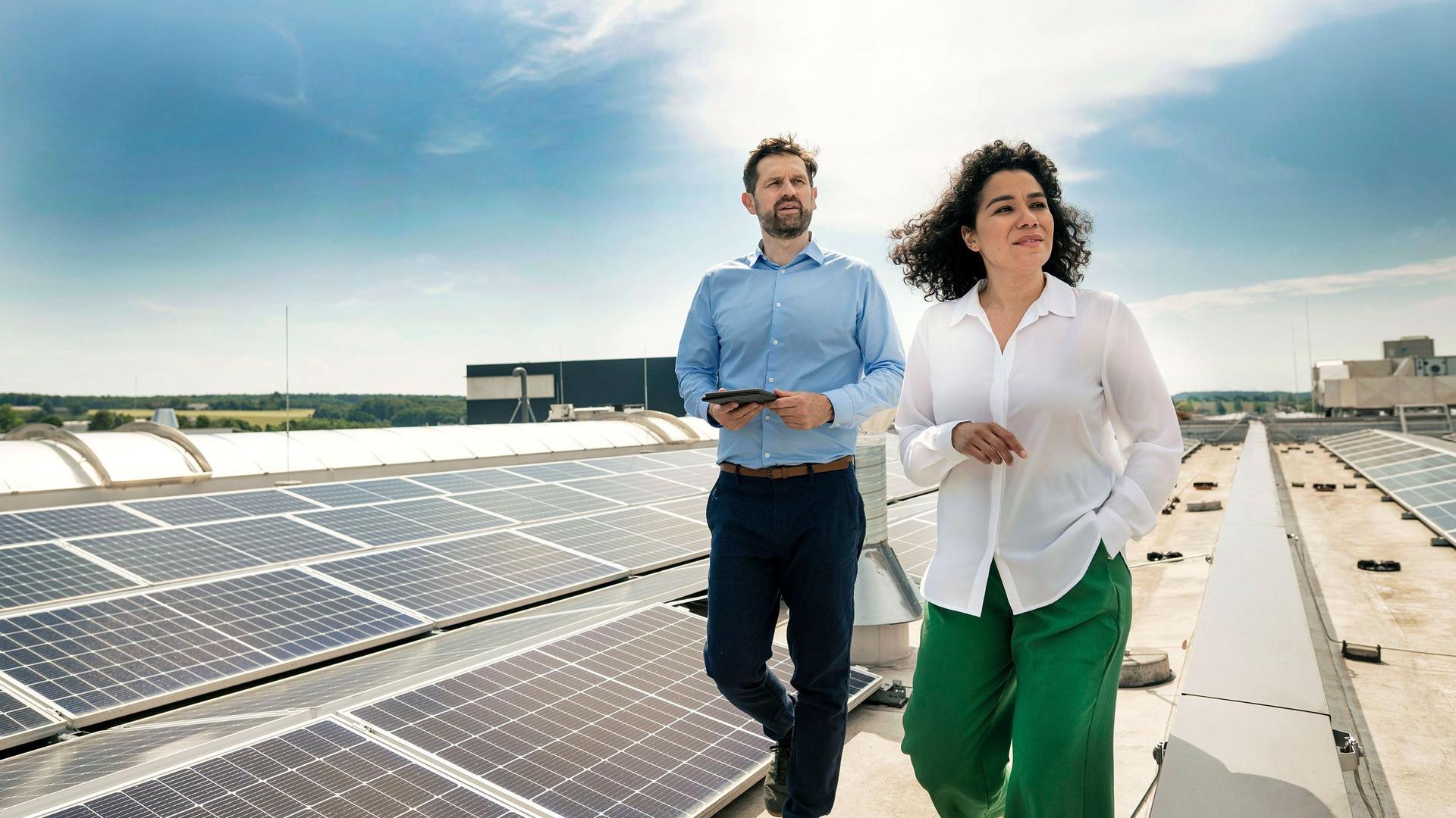 Eine Frau und ein Mann in Businesskleidung laufen bei heiterem Wetter auf dem Dach eines großen Gebäudes, auf welchem eine Photovoltaikanlage installiert ist.