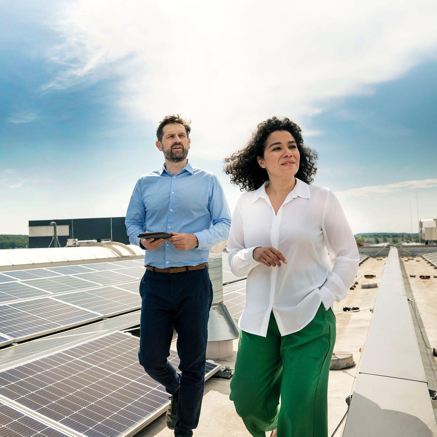 Eine Frau und ein Mann in Businesskleidung laufen bei heiterem Wetter auf dem Dach eines großen Gebäudes, auf welchem eine Photovoltaikanlage installiert ist.