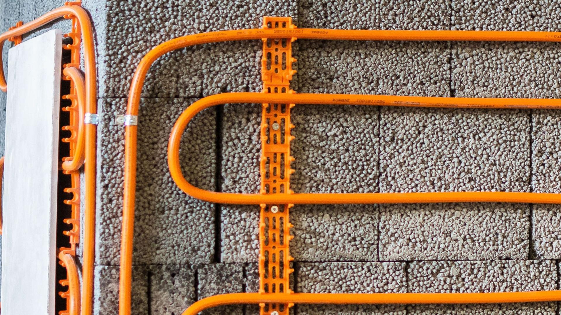 Orangefarbene Kunstoffrohrleitungen für eine Wandheizung mit Halterungen vor unverputzter grauer Mauerwerkswand.