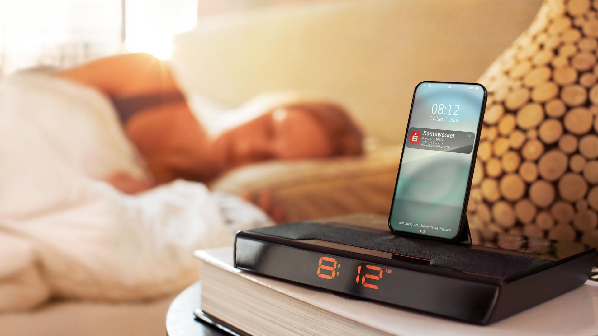 Smartphone und Wecker im Vordergrund, im Hintergrund schlafende Frau im Bett
