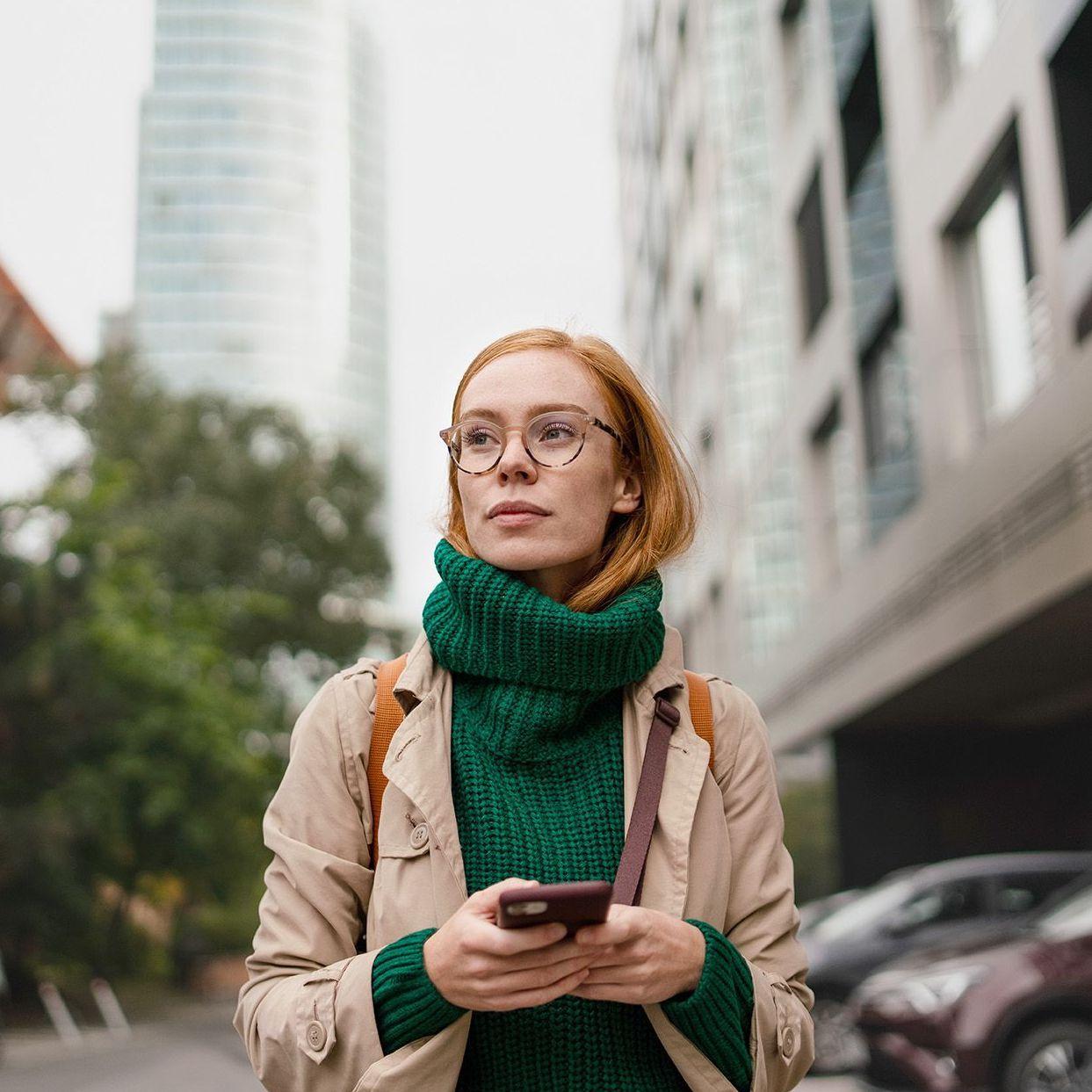 Einer herbstlich gekleidete Frau mit einem Handy in der Hand läuft durch eine Großstadt. Sie schaut nach vorne in die Ferne. Im Hintergrund zeichnen sich Hochhäuser ab.