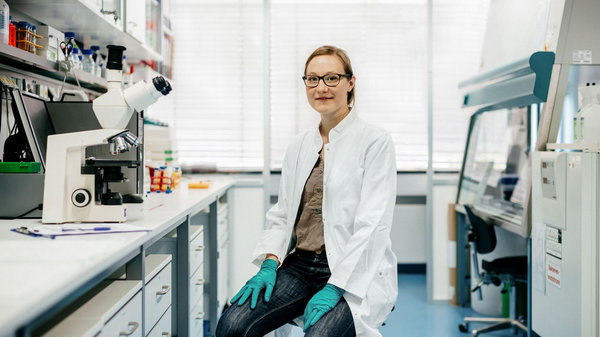 Porträt einer Wissenschaftlerin mit weißem Kittel und Handschuhen auf einem Hocker im modernen Labor. Sie schaut mit einem leichten Lächeln in die Kamera.