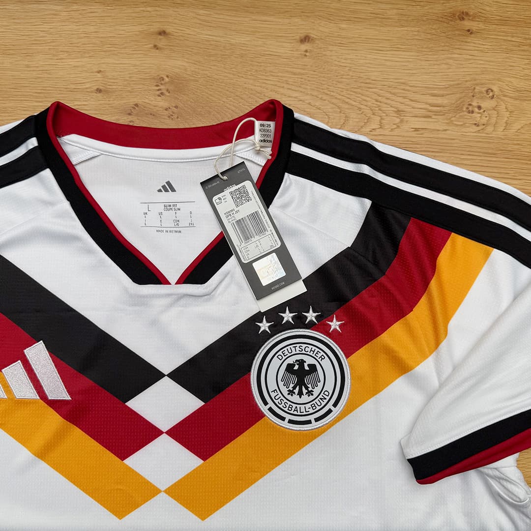 Fußballtrikot in Weiß mit schwarzen, roten und goldenen Streifen, DFB-Logo und Etikett liegt auf einer Holzfläche.