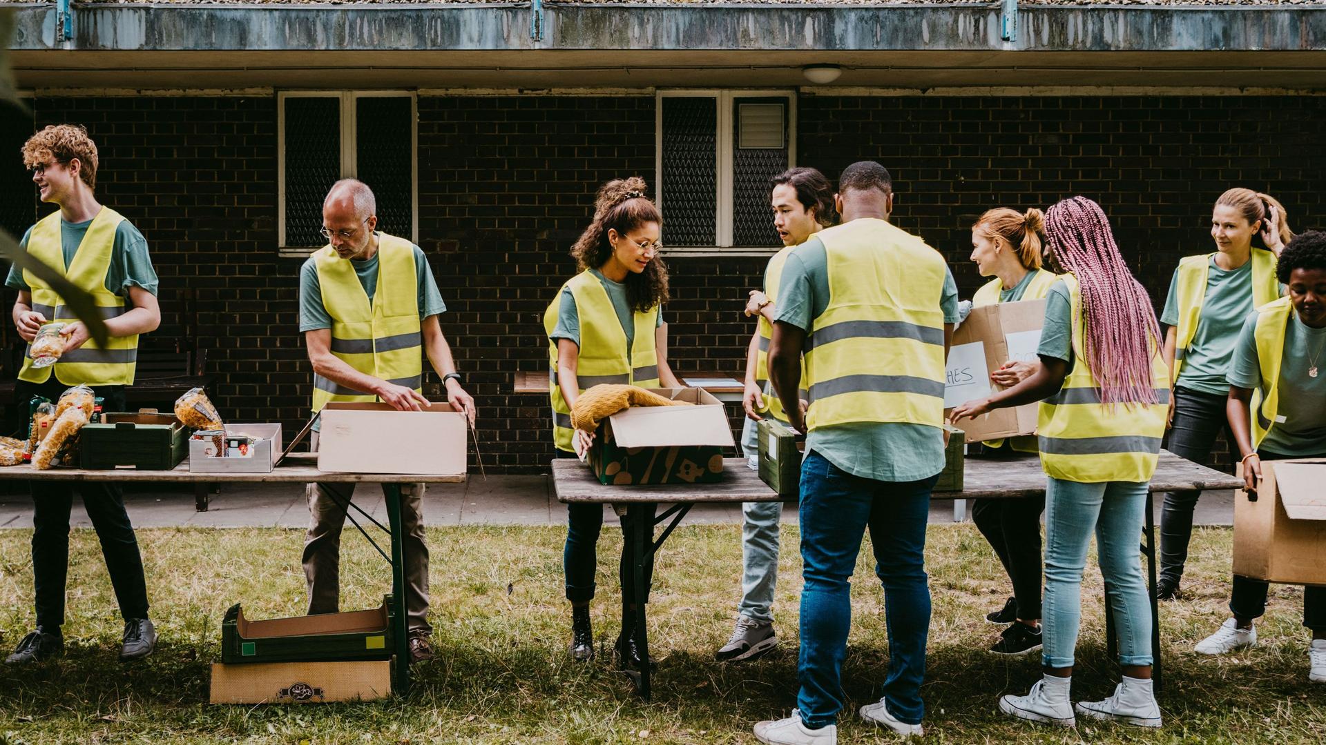 Freiwillige Helfer mit gelben Warnwesten stehen vor Kisten, die auf Tischen stehen, in einem Garten.