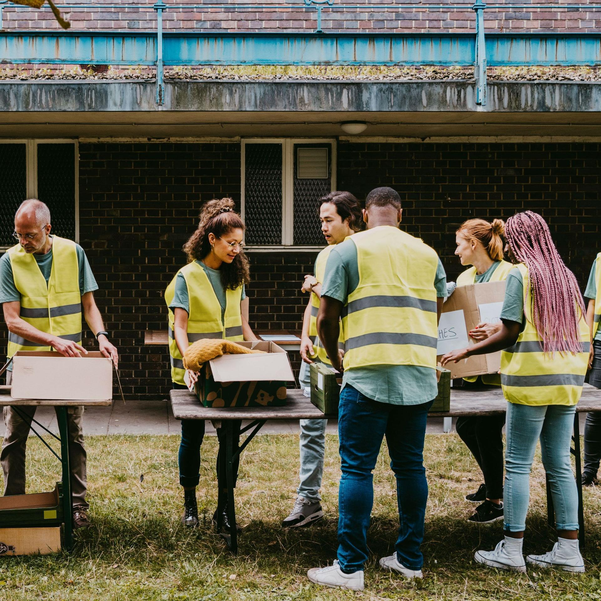 Freiwillige Helfer mit gelben Warnwesten stehen vor Kisten, die auf Tischen stehen, in einem Garten.