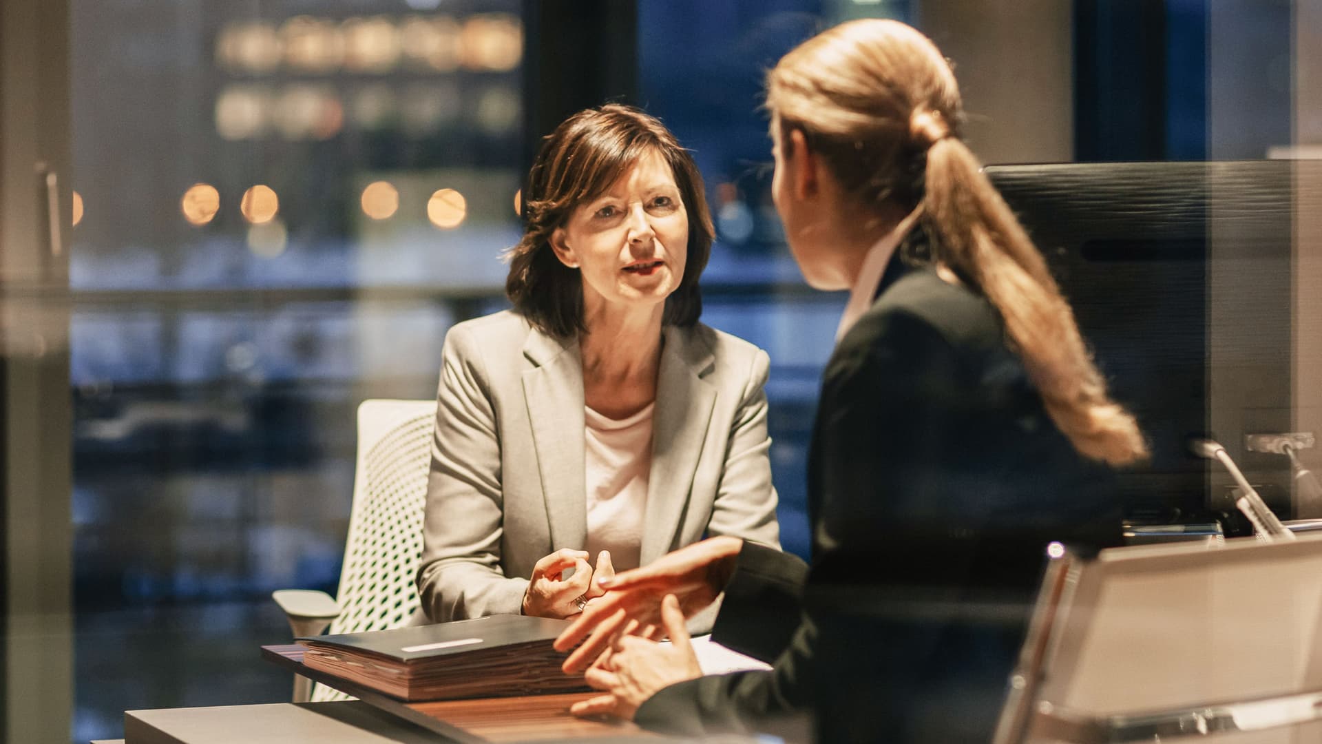 Zwei Frauen in einer Gesprächssituation in einem Büro mit großer Fensterfront. Draußen ist es dunkel.