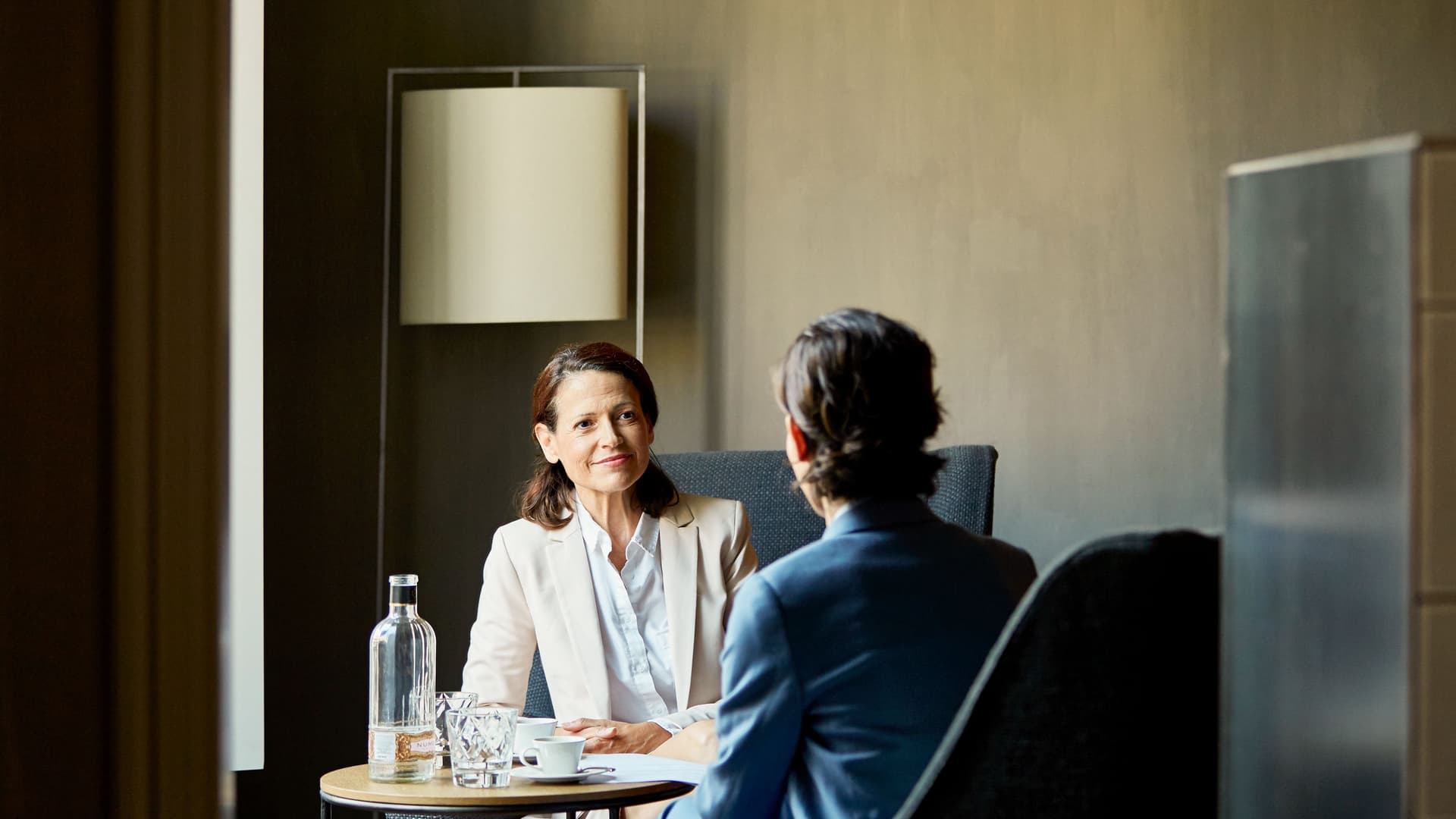 Eine Frau in Business-Kleidung spricht mit einem Mann an einem Tisch in einem Hotelzimmer.