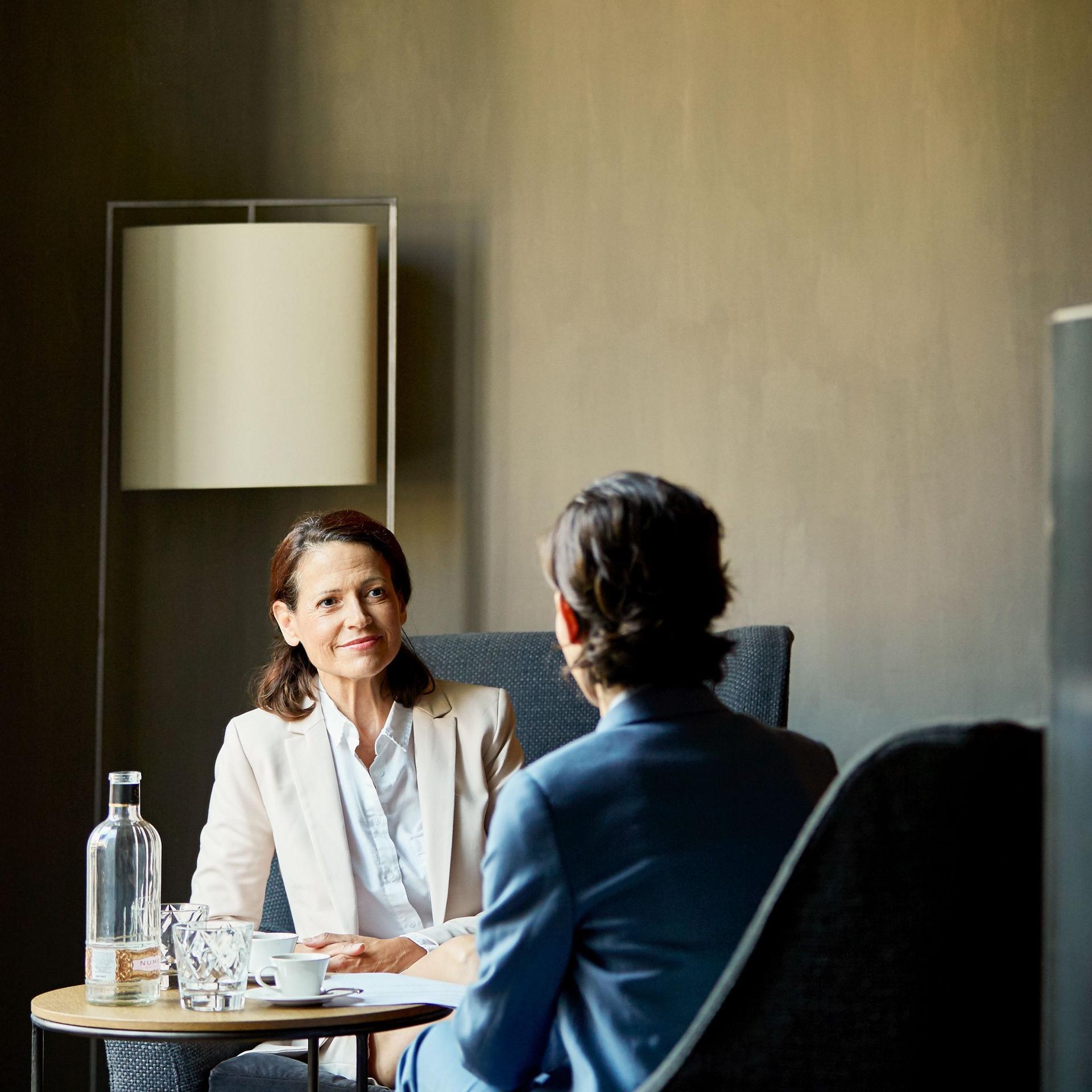 Eine Frau in Business-Kleidung spricht mit einem Mann an einem Tisch in einem Hotelzimmer.