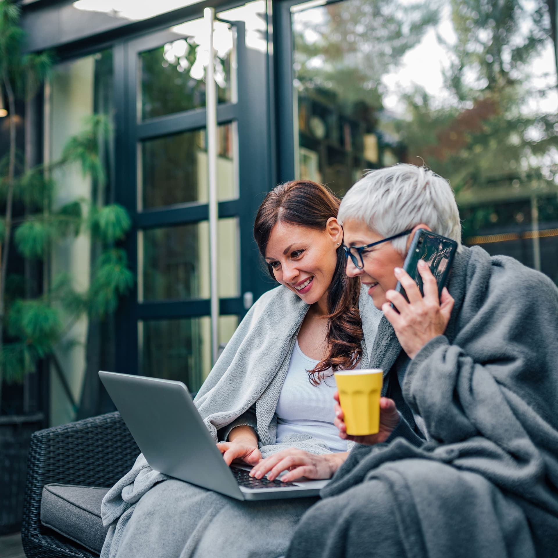 Zwei Frauen sitzen auf einer Terasse. Die eine hat ein Handy am Ohr, die andere einen Laptop auf dem Schoß.
