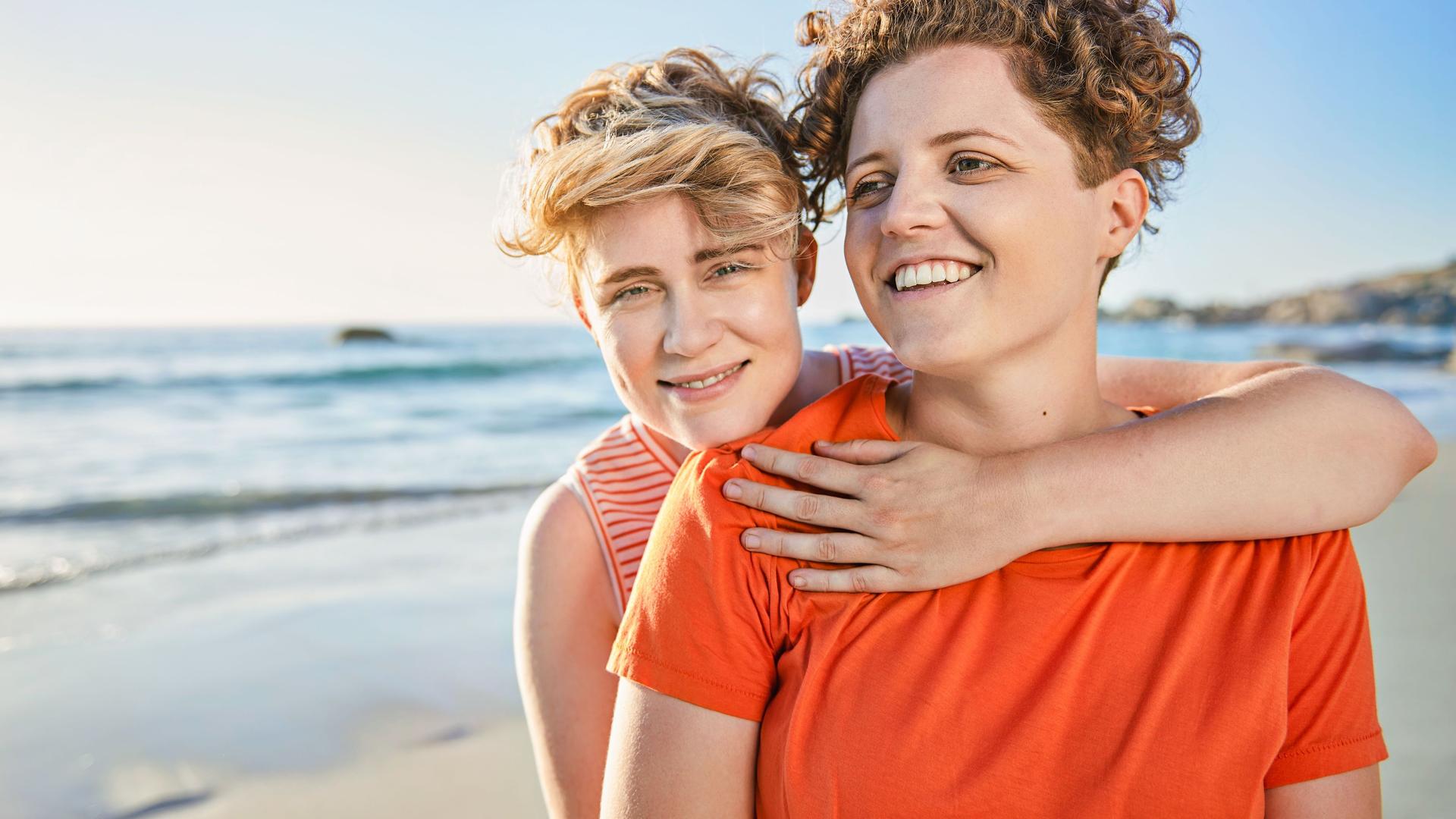 Zwei junge Frauen umarmen sich am Strand. Sie lächeln beide in die Kamera.