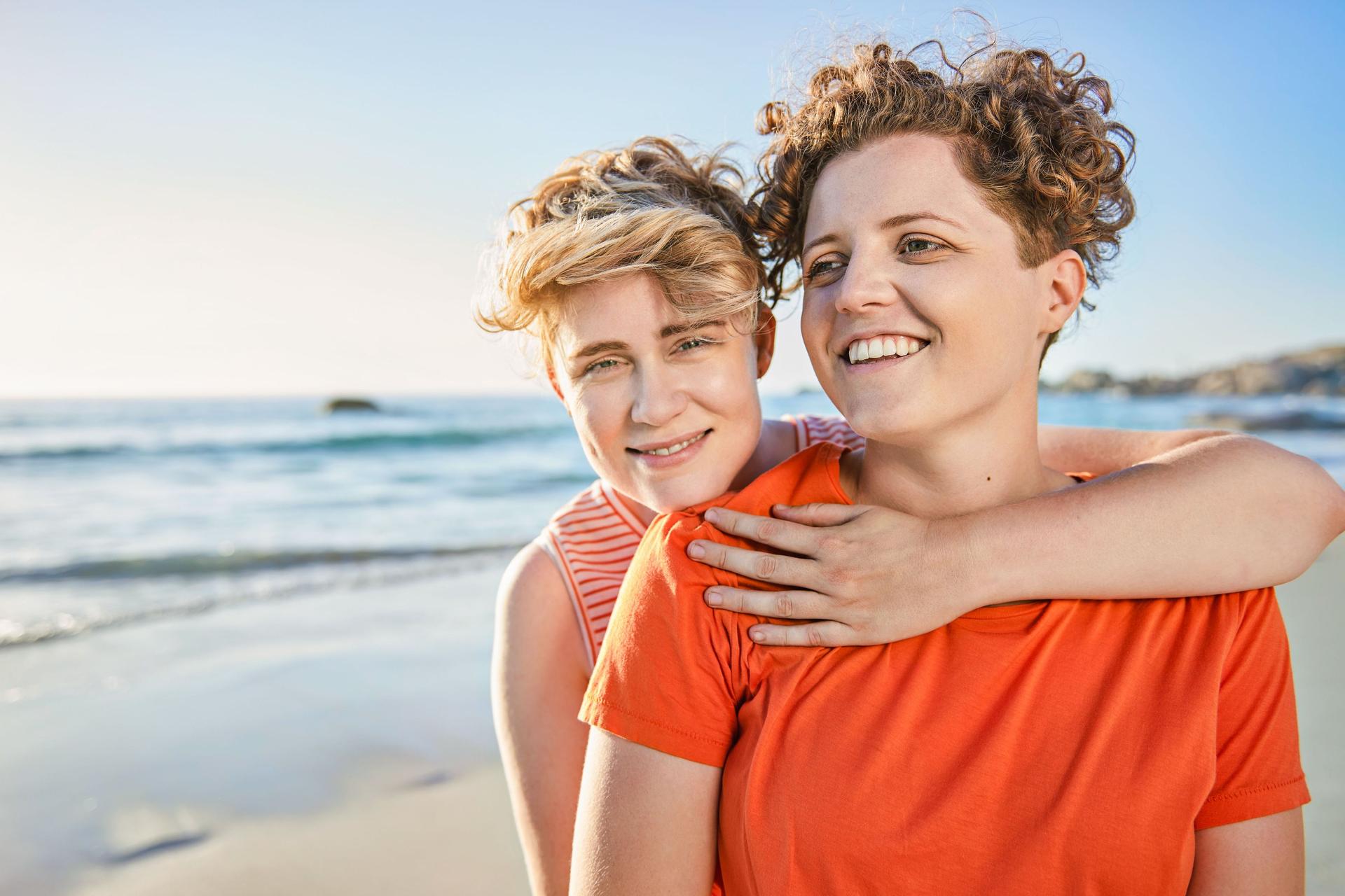 Zwei junge Frauen umarmen sich am Strand. Sie lächeln beide in die Kamera.