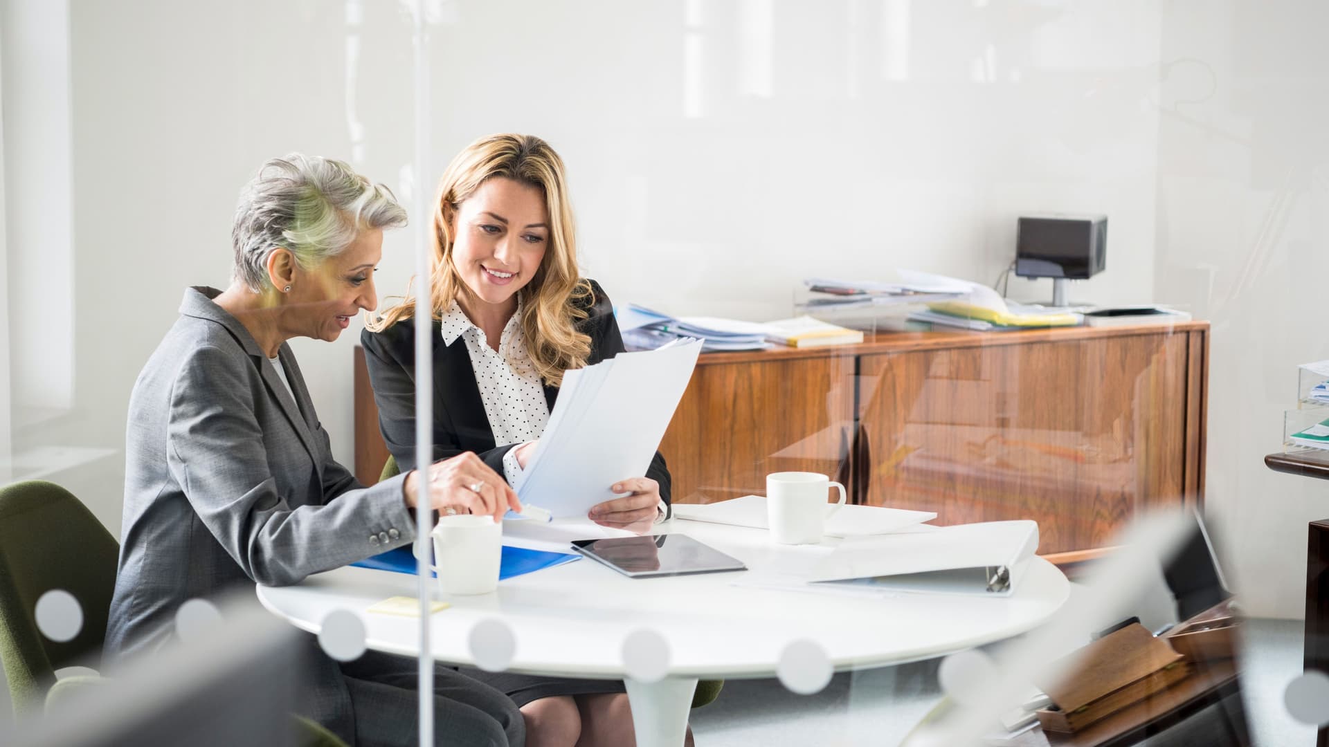 Zwei Frauen sitzen an einem runden Tisch in einem verglasten Büro und schauen auf Papierdokumente in ihren Händen.