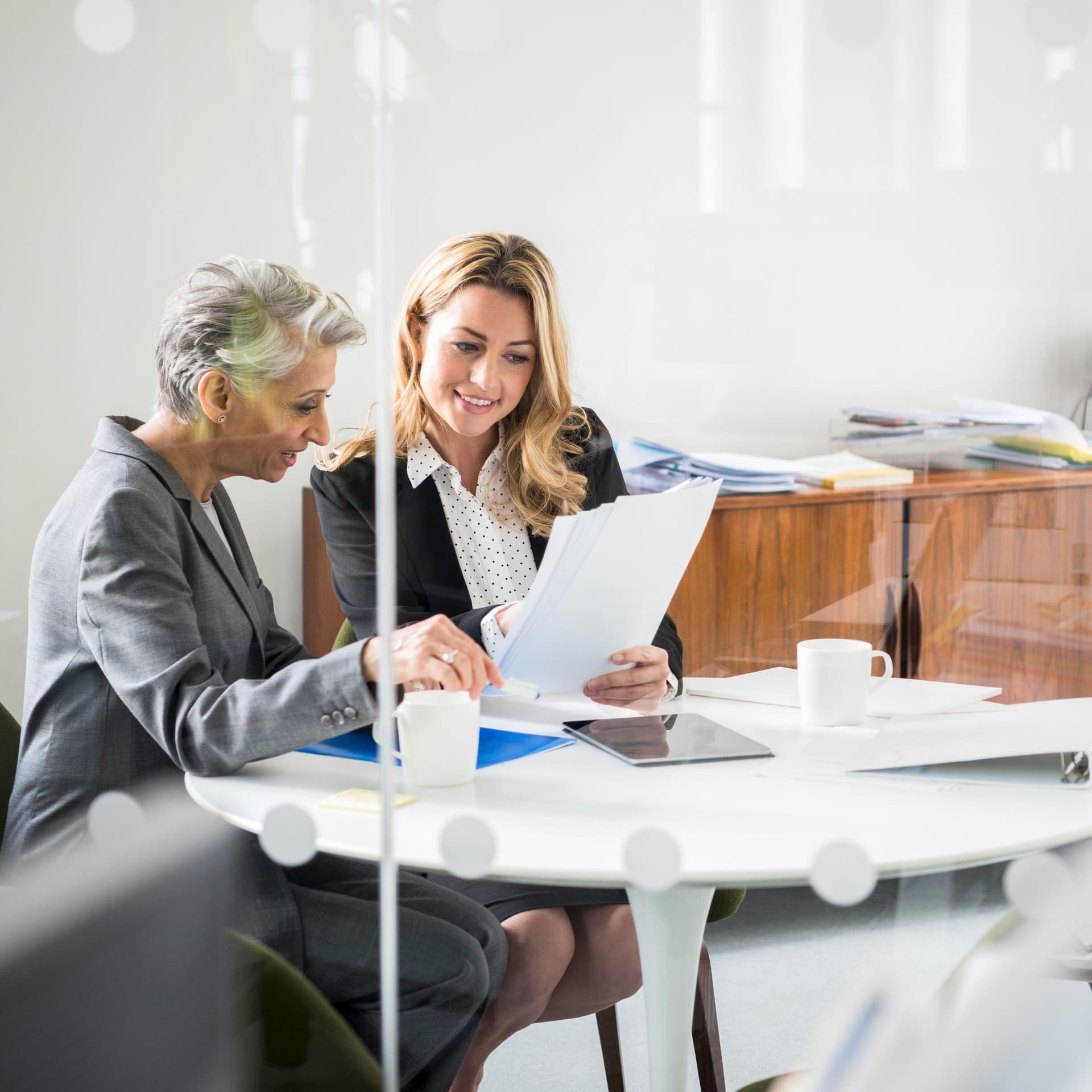 Zwei Frauen sitzen an einem runden Tisch in einem verglasten Büro und schauen auf Papierdokumente in ihren Händen.