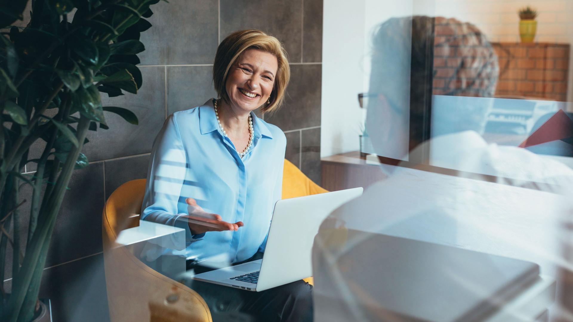 Eine Geschäftsfrau in blauer Bluse unterhält sich angeregt mit einem Geschäftspartner und lächelt. Sie lacht und hat einen Laptop auf dem Schoß.