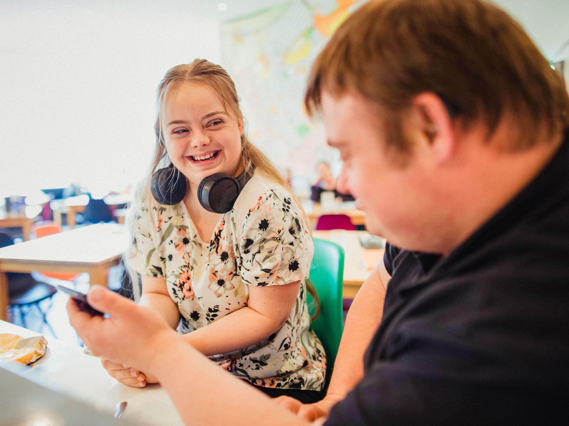 Zwei junge Menschen sitzen in einem Klassenzimmer, unterhalten sich lachend und schauen gemeinsam in ein Smartphone.