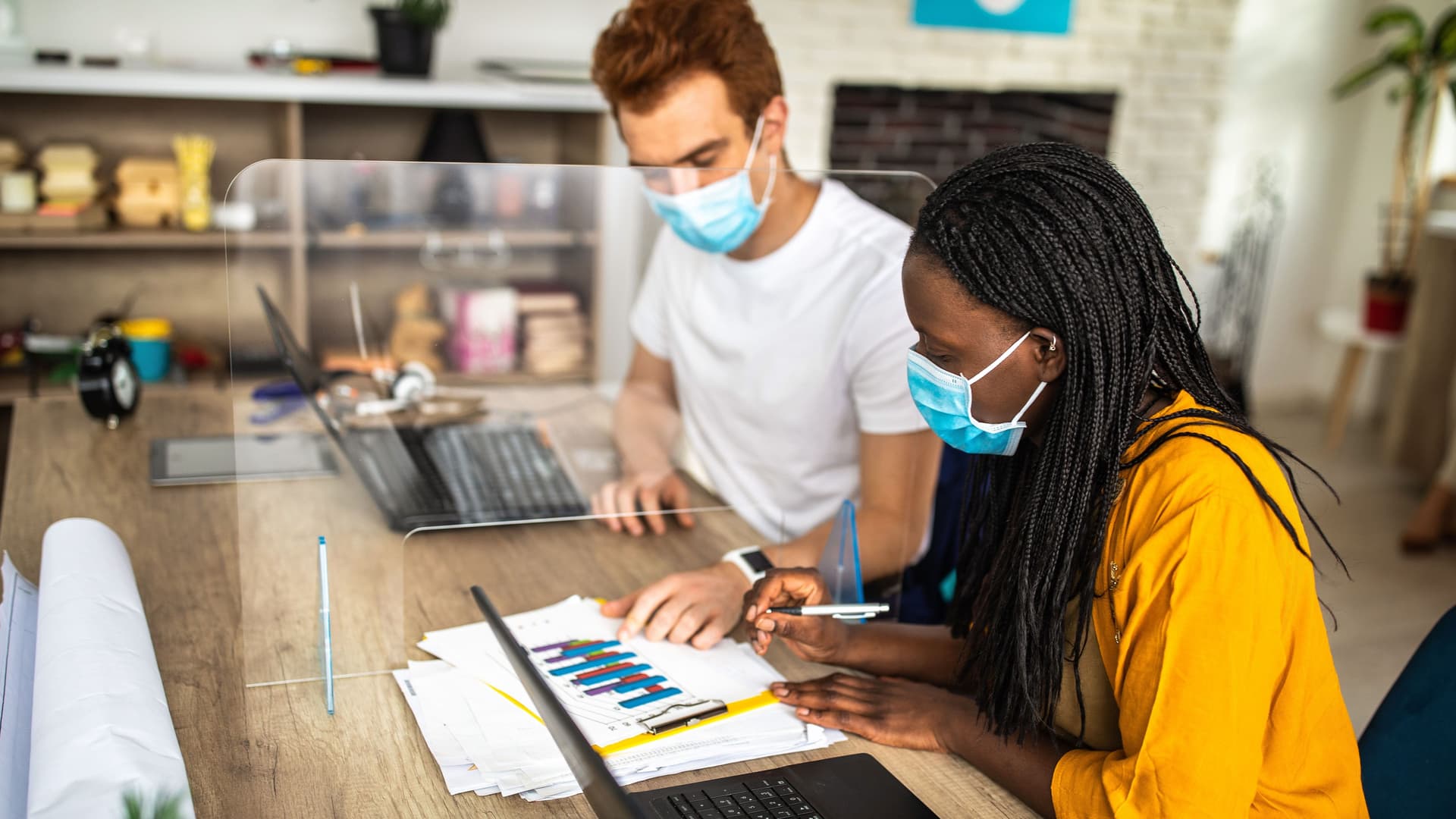 Eine Frau und ein Mann sitzen am Schreibtisch und arbeiten gemeinsam an Diagrammen während sie eine Maske tragen. Eine Plexiglasscheibe steht zwischen ihnen.