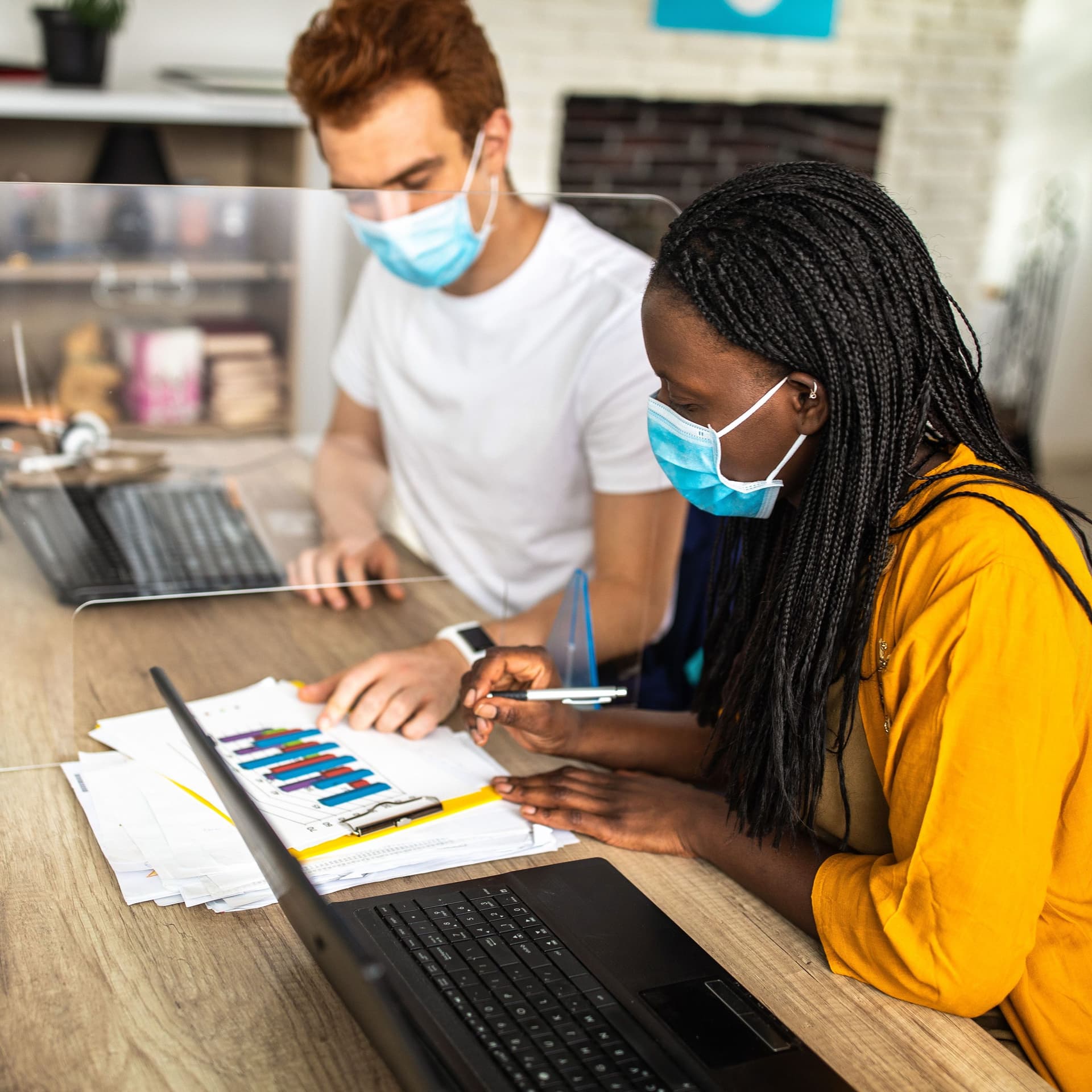 Eine Frau und ein Mann sitzen am Schreibtisch und arbeiten gemeinsam an Diagrammen während sie eine Maske tragen. Eine Plexiglasscheibe steht zwischen ihnen.
