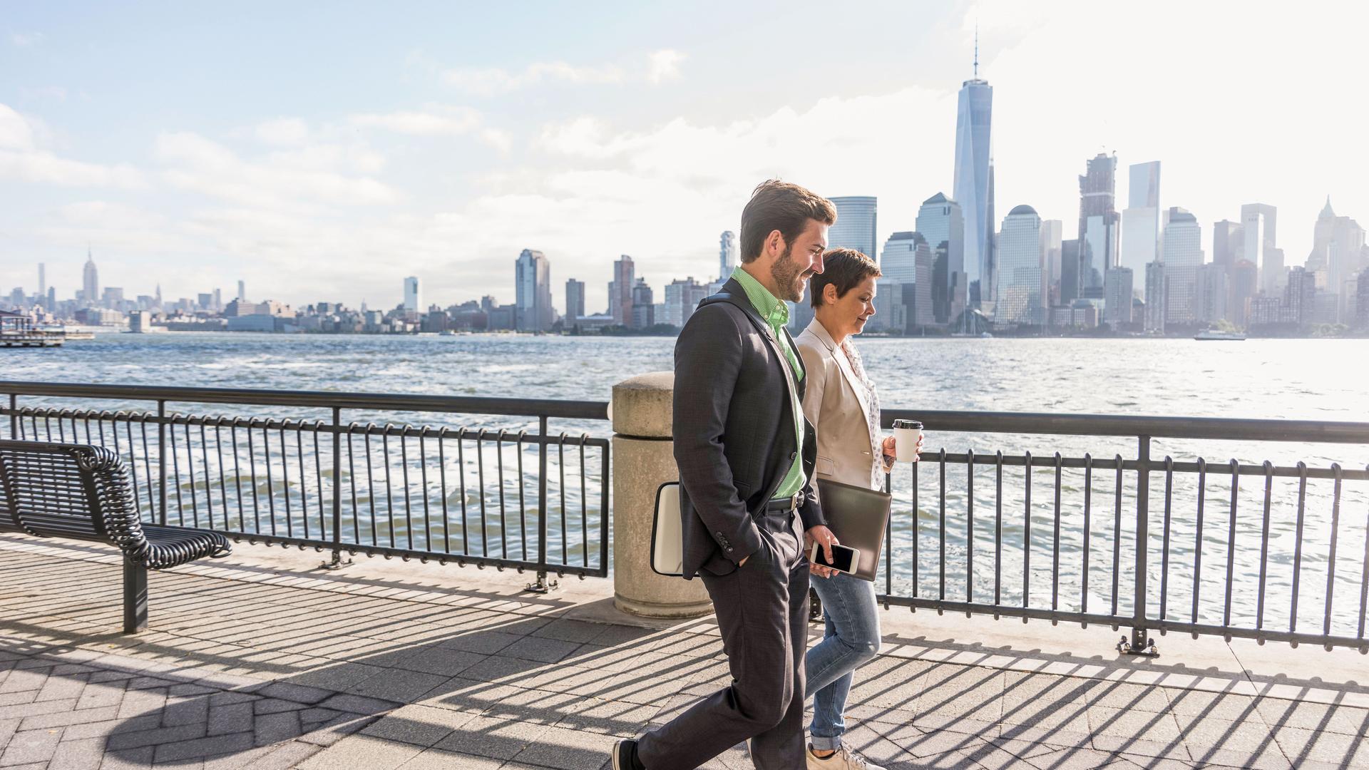 Eine Frau und ein Mann laufen nebeneinander am Wasser entlang. Sie sind förmlich gekleidet. Auf der anderen Flussseite ist die Skyline von Manhattan zu sehen.