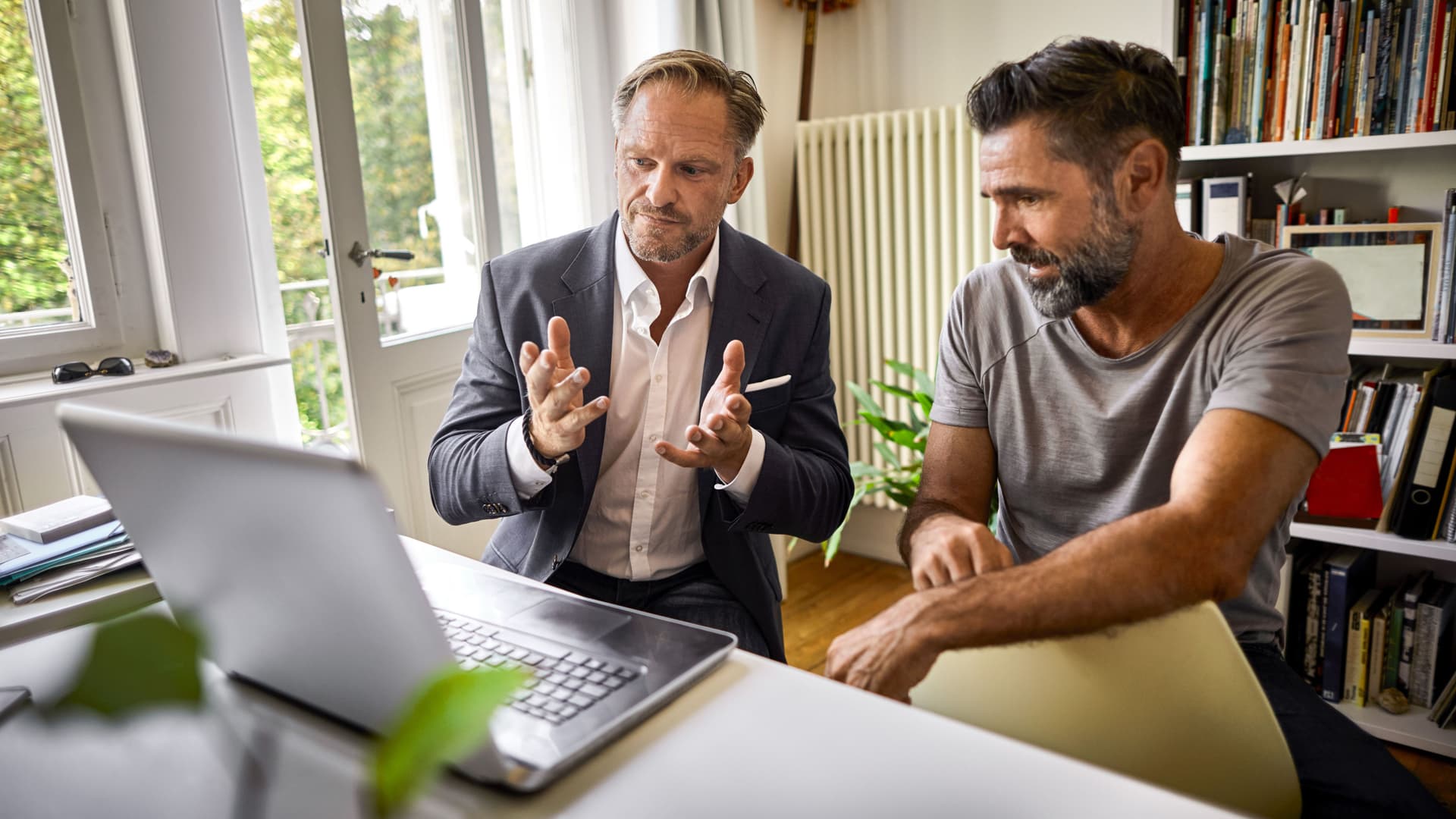 Zwei Männer sitzen in einer Altbauwohnung vor einem Laptop und gestikulieren Richtung Laptop.