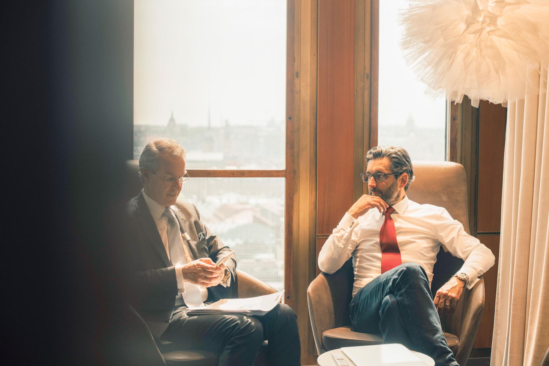 Zwei Männer in Business Kleidung sitzen diskutierend vor einem großen Fenster.
