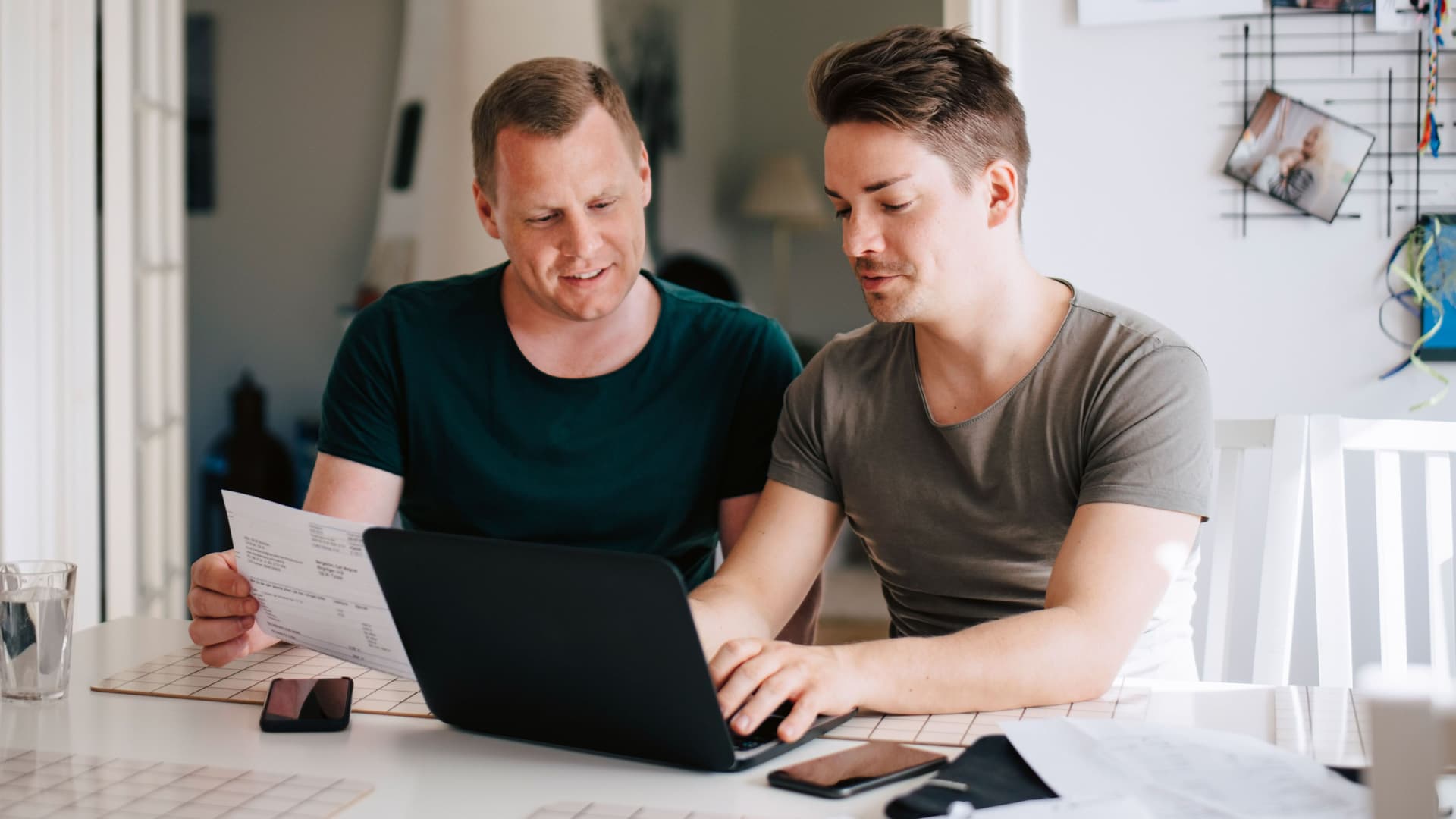 Zwei Männer am Küchentisch mit Laptop zahlen Rechnungen online.