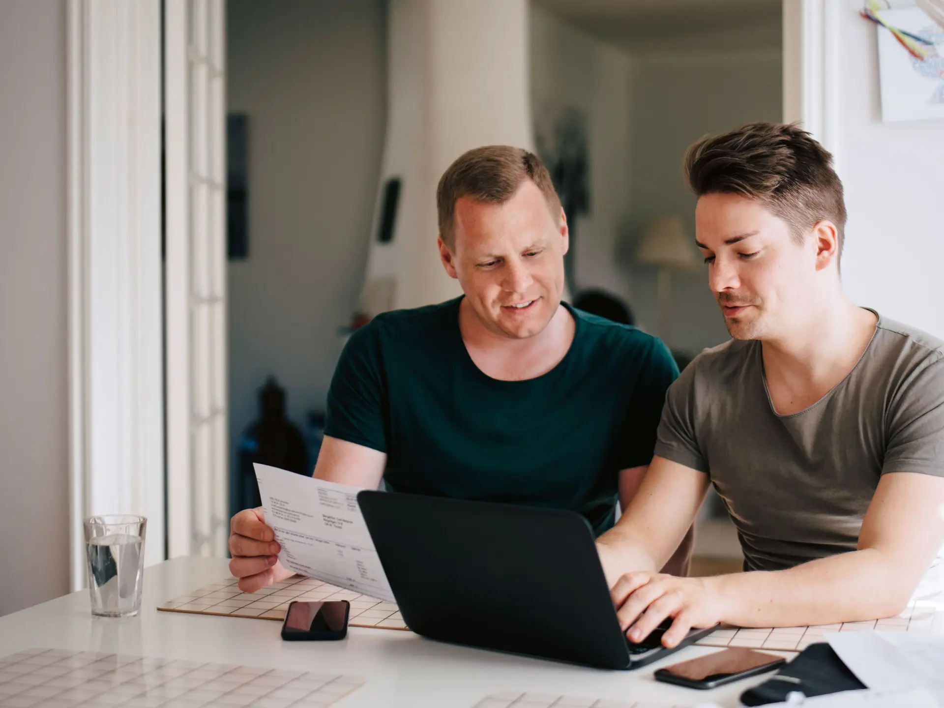 Zwei Männer am Küchentisch mit Laptop zahlen Rechnungen online.