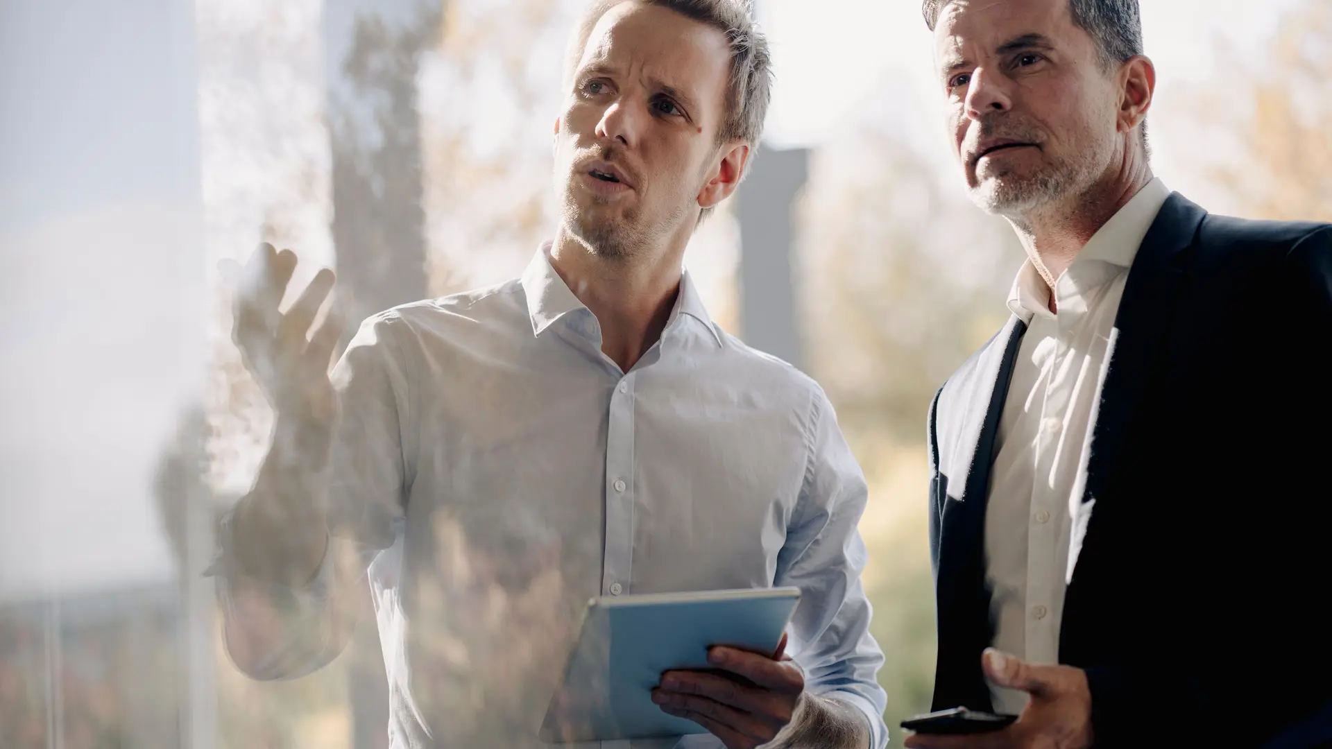 Zwei Männer in Business-Kleidung unterhalten sich. Ein Mann gestikuliert und hält Papiere in der Hand, der andere Mann hält ein Smartphone. Beide blicken durch eine Glasscheibe nach oben.