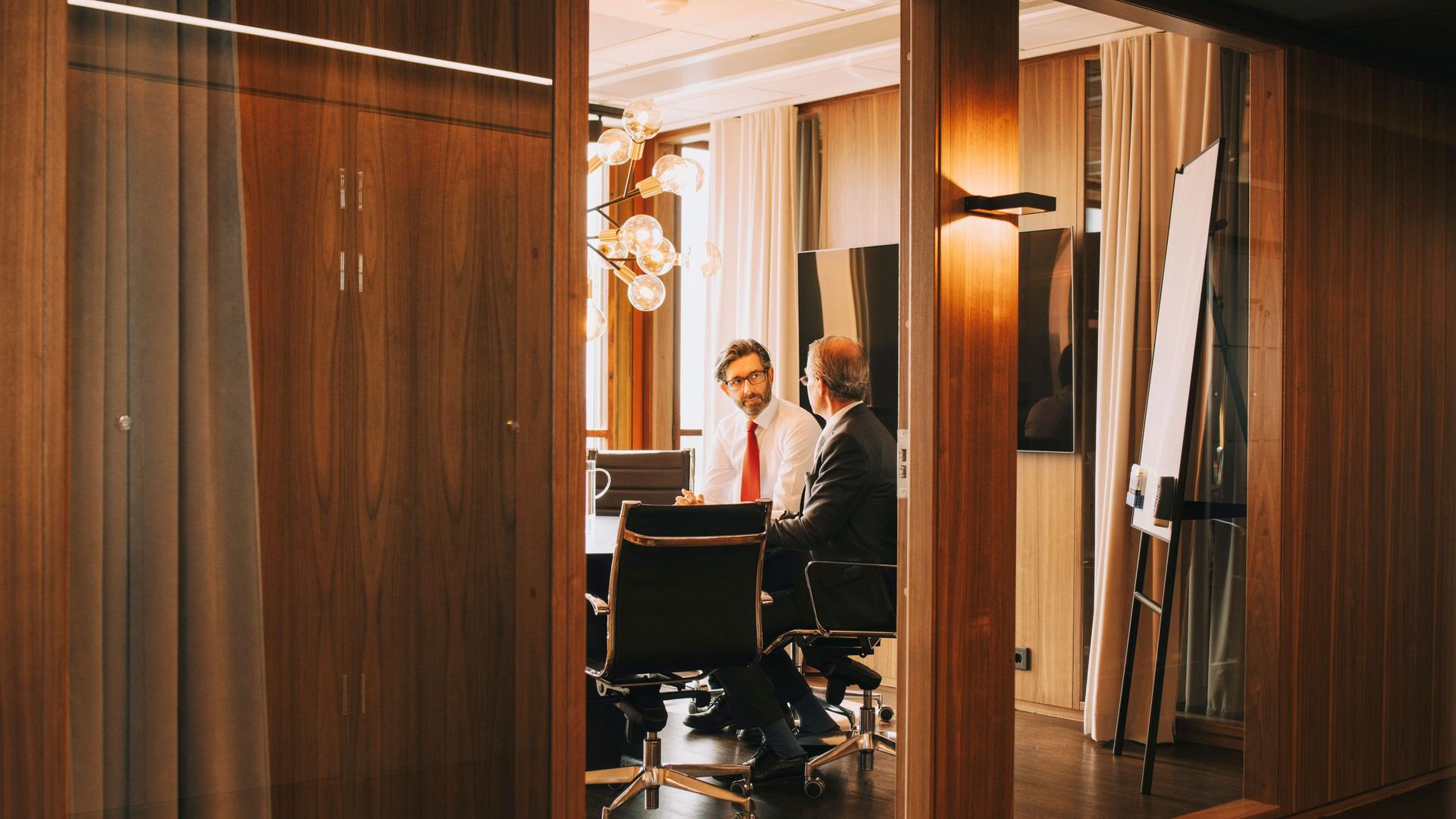 Zwei Männer mittleren Alters sitzen in einem Meeting Raum. Hinter ihnen ist ein großer Bildschirm und ein Whiteboard zu sehen.