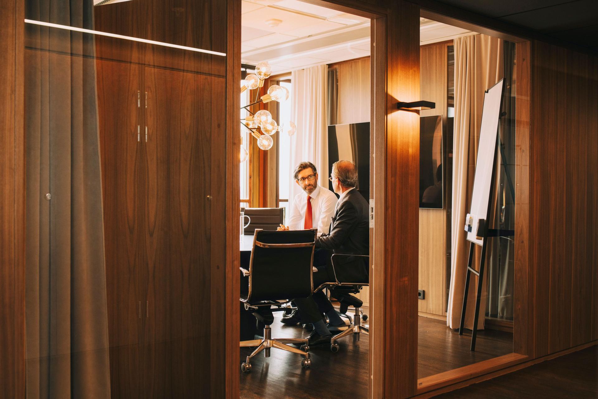 Zwei Männer mittleren Alters sitzen in einem Meeting Raum. Hinter ihnen ist ein großer Bildschirm und ein Whiteboard zu sehen.