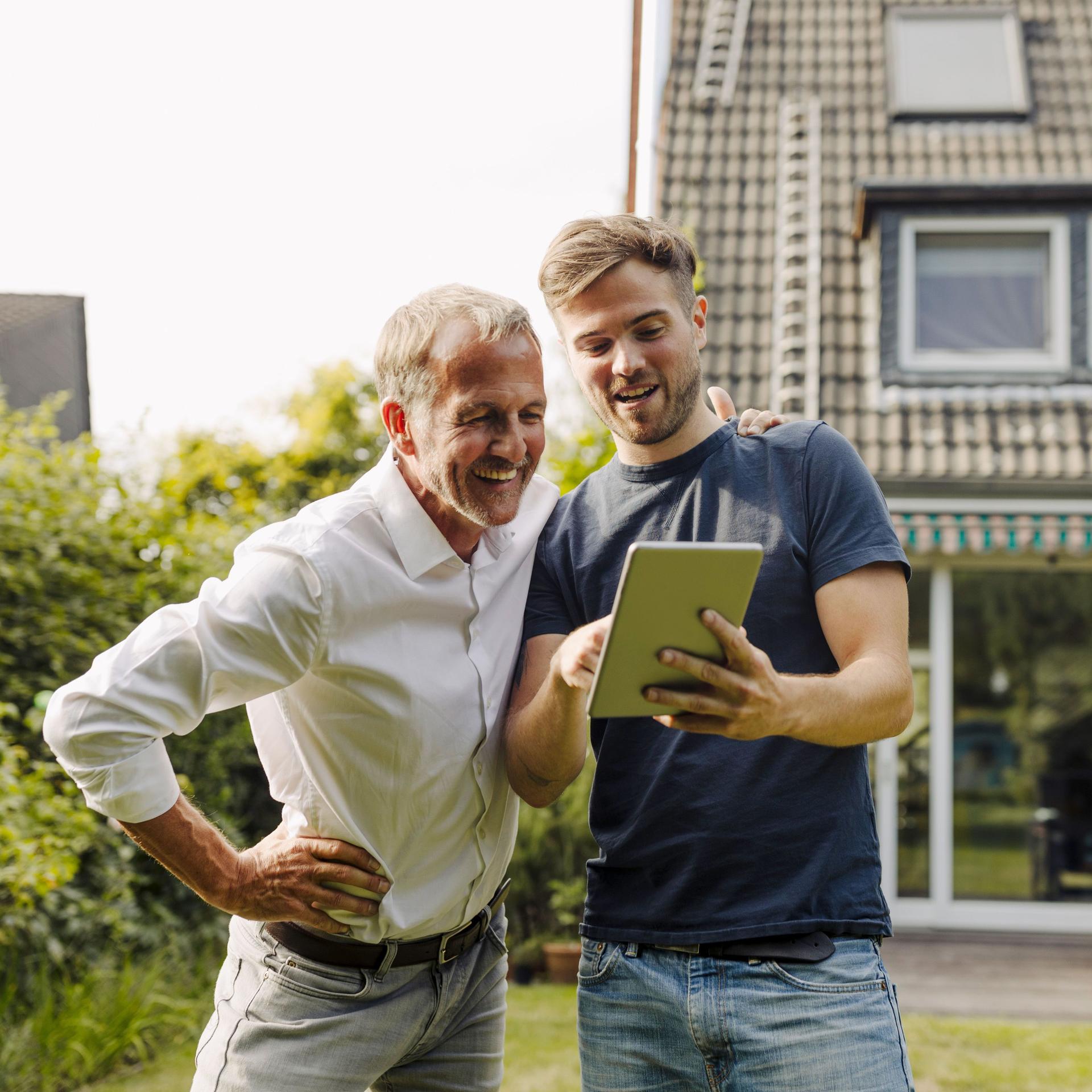 Zwei Männer stehen im Garten mit einem Tablet in der Hand. Sie schauen auf das Tablet. Im Hintergrund ist ein Einfamilienhaus zu sehen.