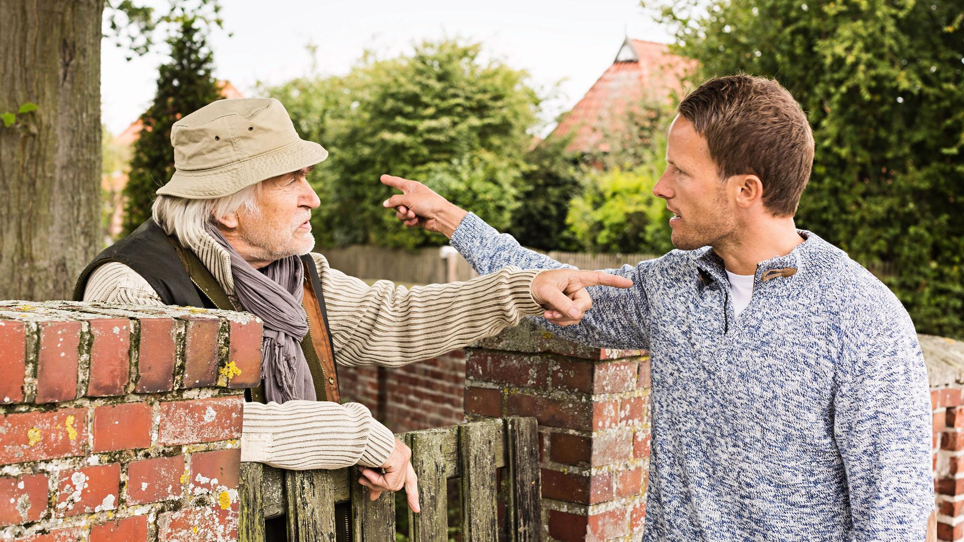 Ein älterer Mann und ein weiterer erwachsener Mann streiten sich am Gartentor. Sie drohen sich mit Gesten.