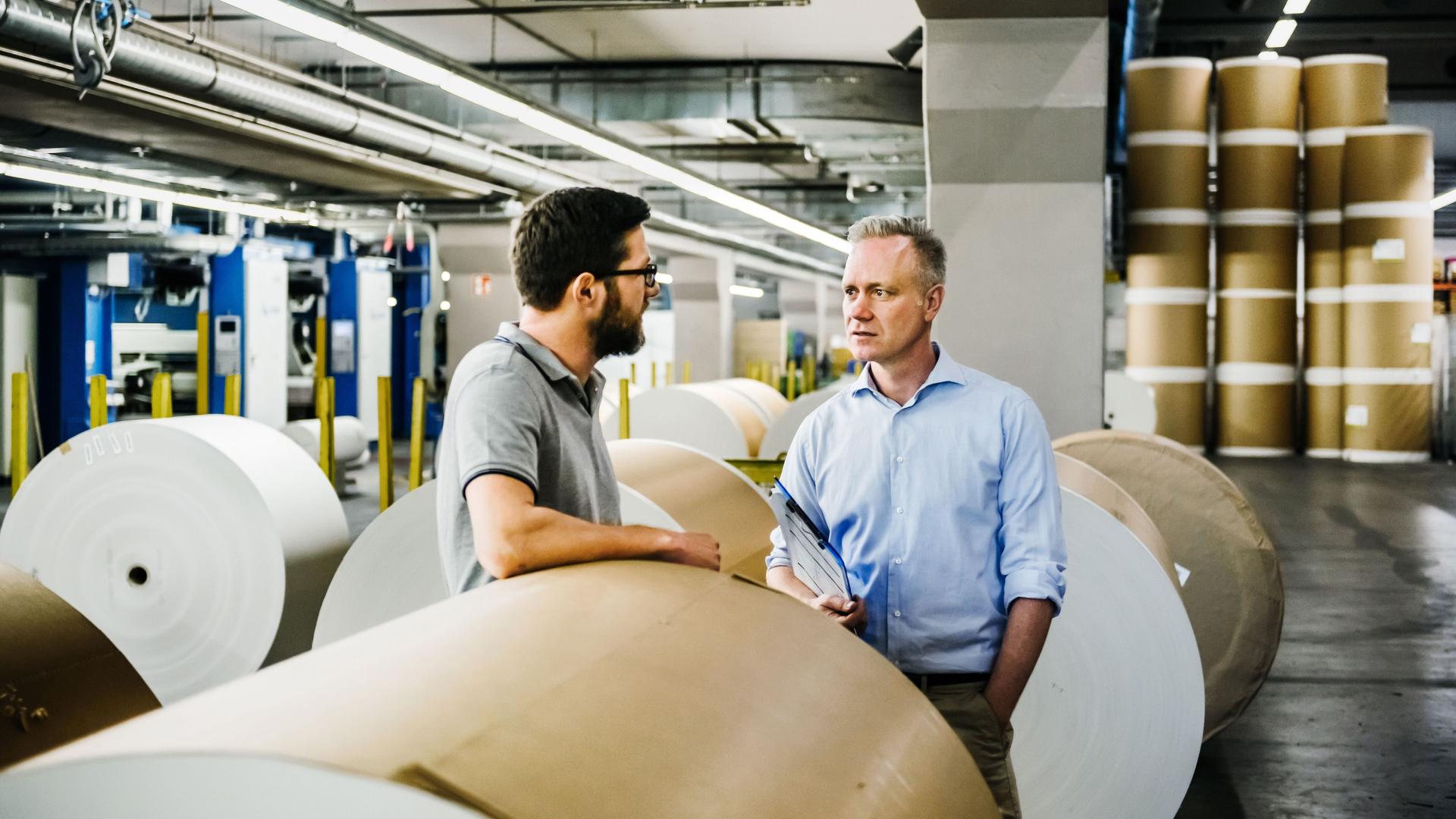 Zwei Männer unterhalten sich in einer Papierfabrik. Im Hintergrund stehen Papierrollen.
