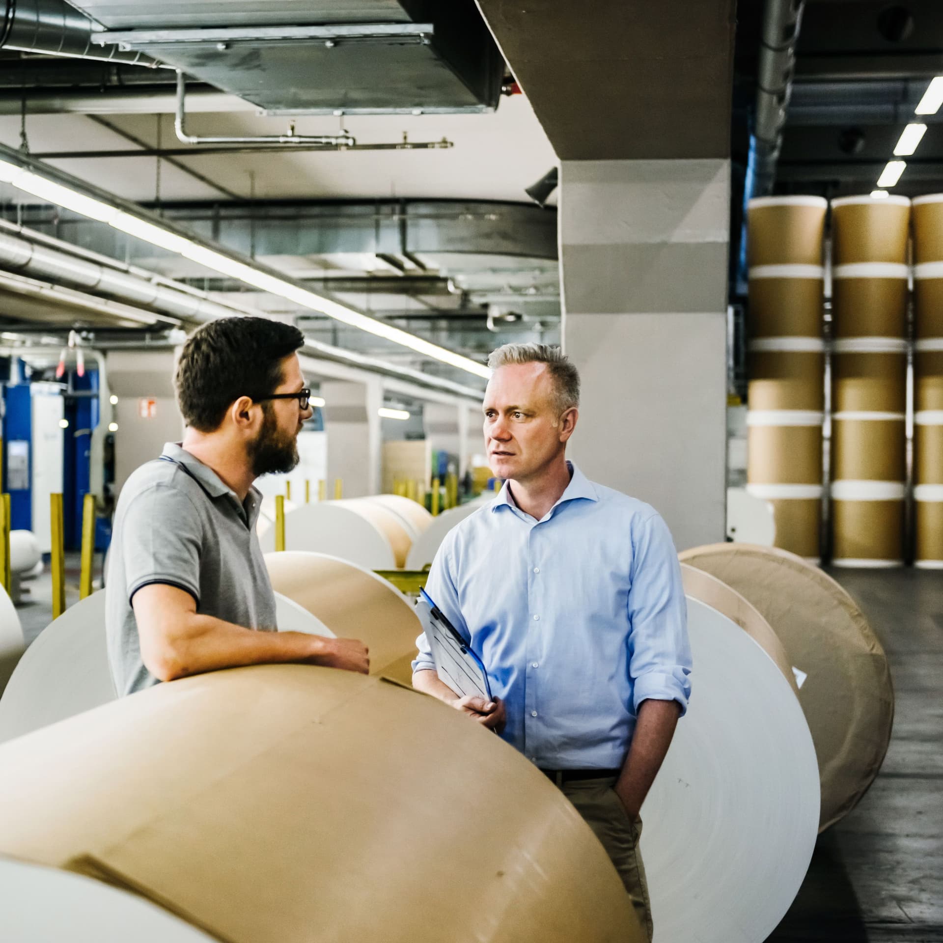 Zwei Männer unterhalten sich in einer Papierfabrik. Im Hintergrund stehen Papierrollen.