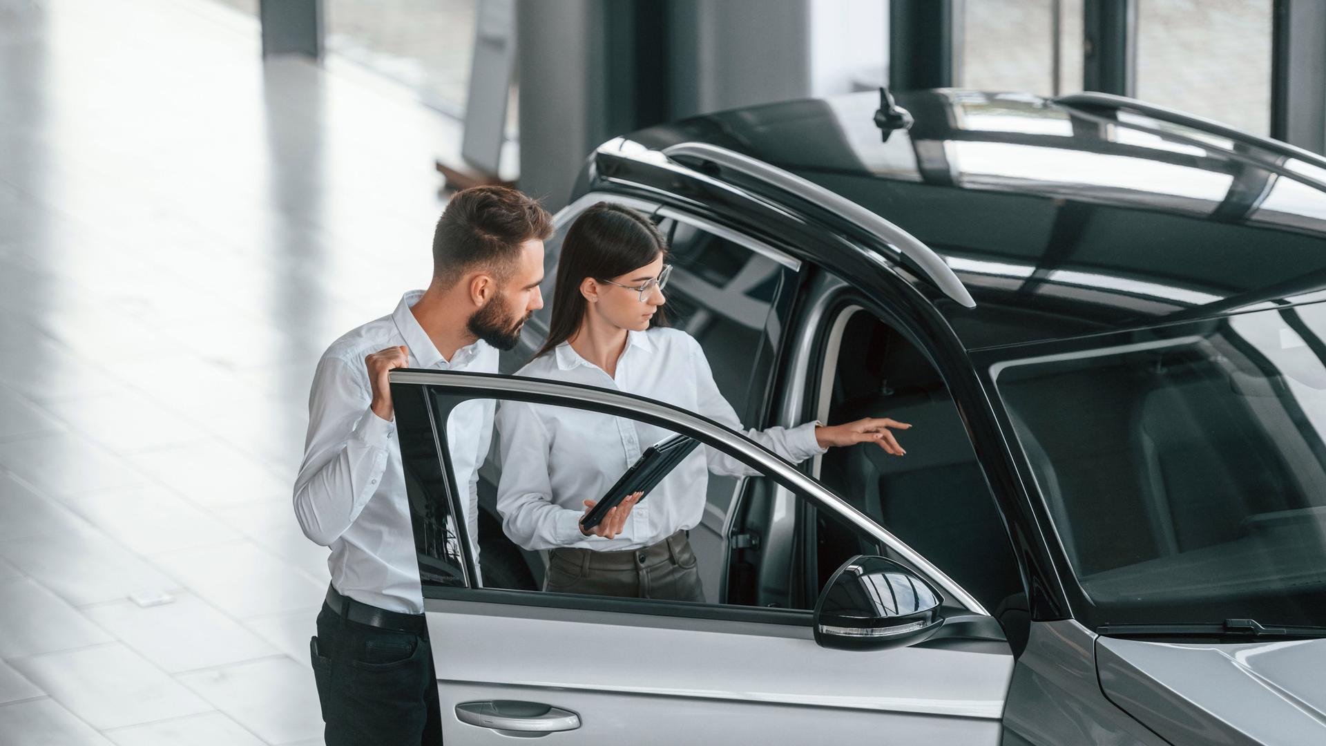 Ein Mann und eine Frau in weißen Hemden stehen an der offenen Tür eines SUVs. Die Frau hält in der einen Hand ein Tablet und zeigt mit der anderen in das Fahrzeuginnere.