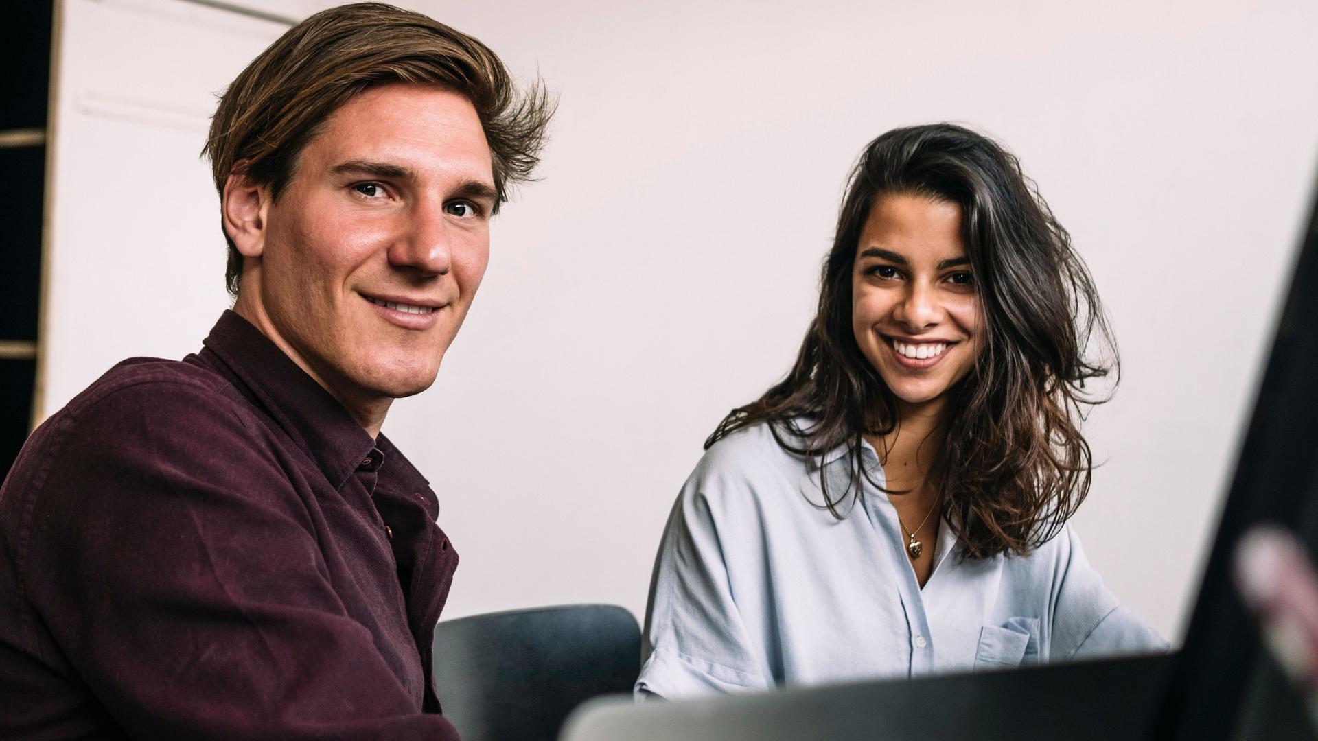 Porträt zweier Studenten, die vor einem Computer sitzen und lächelnd in die Kamera schauen.