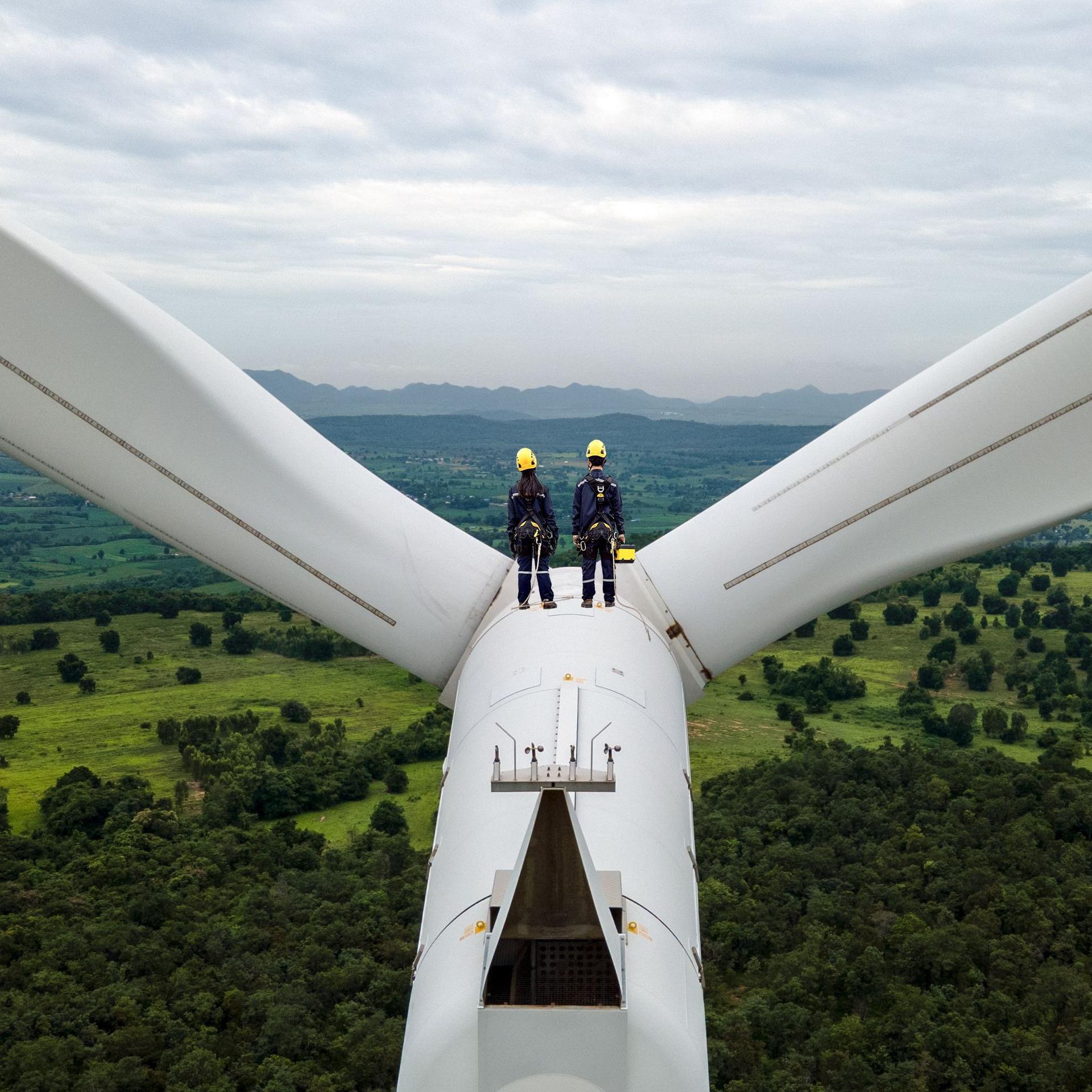 Zwei Elektroingenieure mit Schutzausrüstung stehen oben auf einem Windrad zwischen den Windblättern. Sie schauen in die Landschaft.