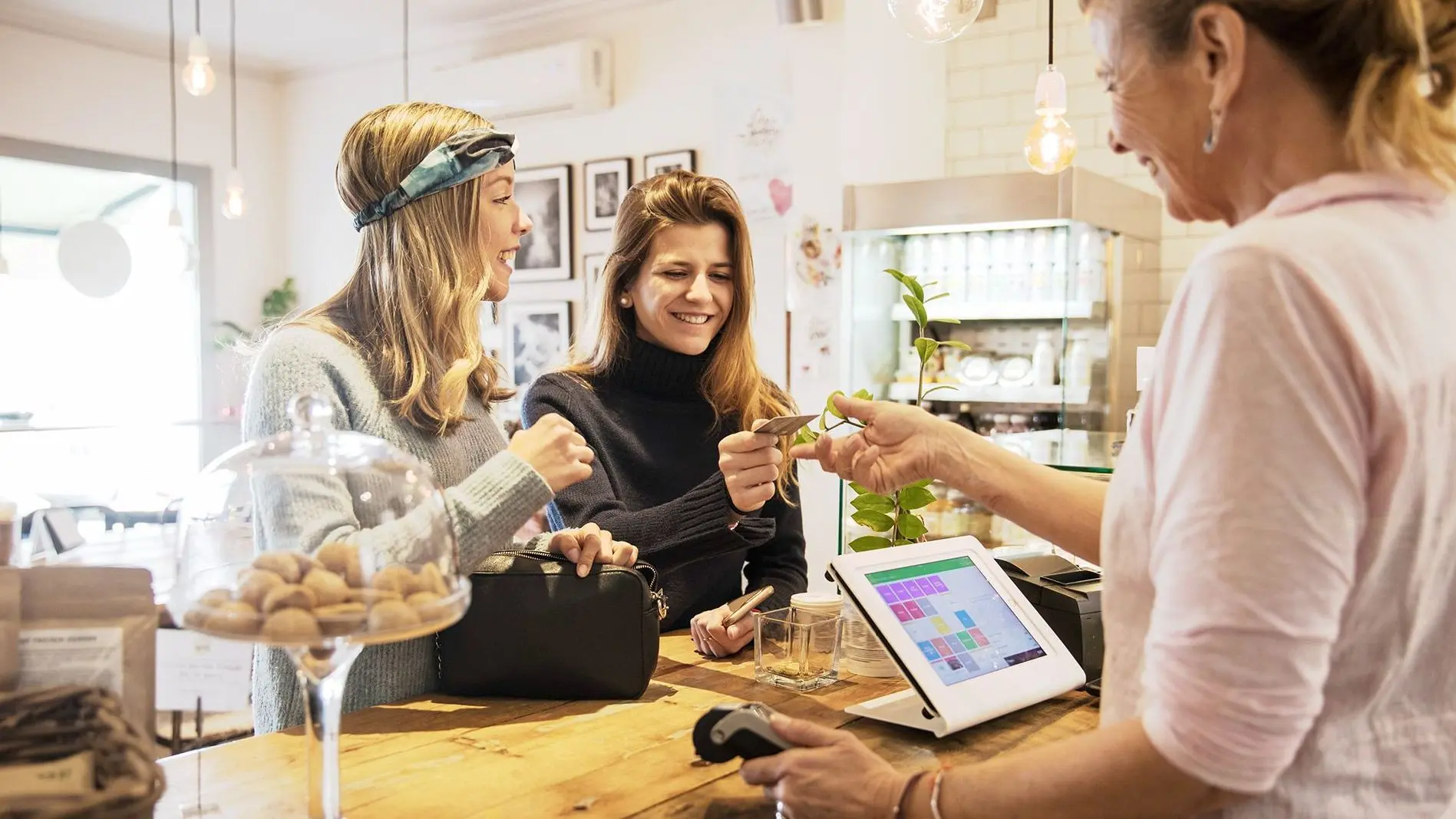 Zwei Freundinnen im Teenageralter, die an der Theke eines Cafés stehen und der Verkäuferin eine Kreditkarte zum Bezahlen geben. Sie lachen ausgelassen.