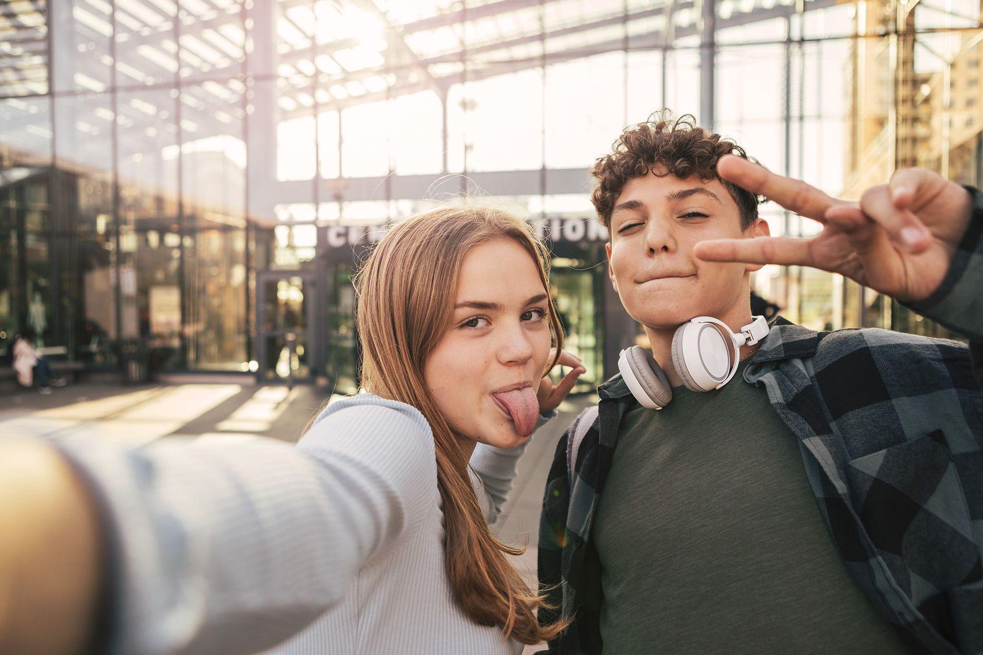 Zwei Jugendliche machen mit einem Smartphone ein Selfie vor einem Glasgebäude.